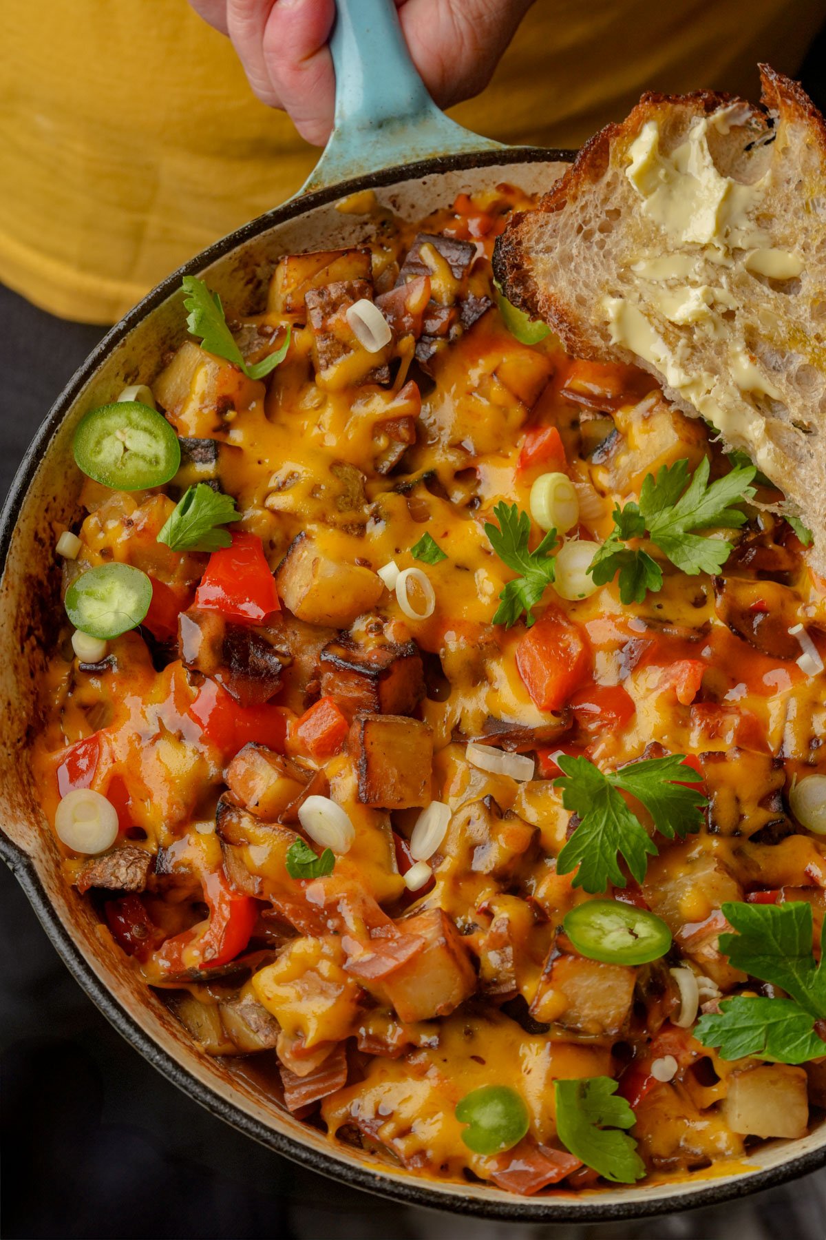 A skillet filled with cheesy vegan breakfast hash made of diced potatoes, red bell peppers, onions, veggie bacon, and herbs, topped with green onions; a slice of buttered bread rests on the side.