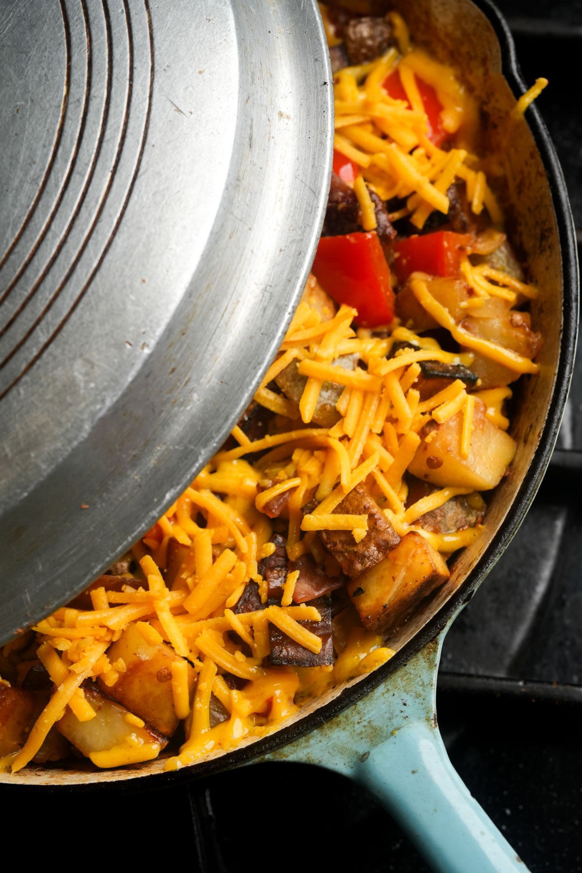 A skillet with vegan breakfast hash and shredded cheddar cheese is partially covered by a metal lid.