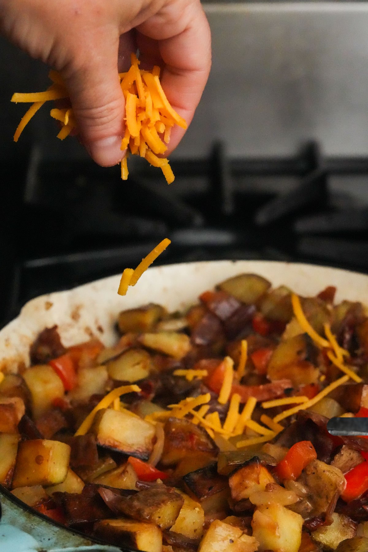 A hand sprinkles shredded cheddar cheese over a skillet containing cooked potatoes, peppers, and onions on a stove.