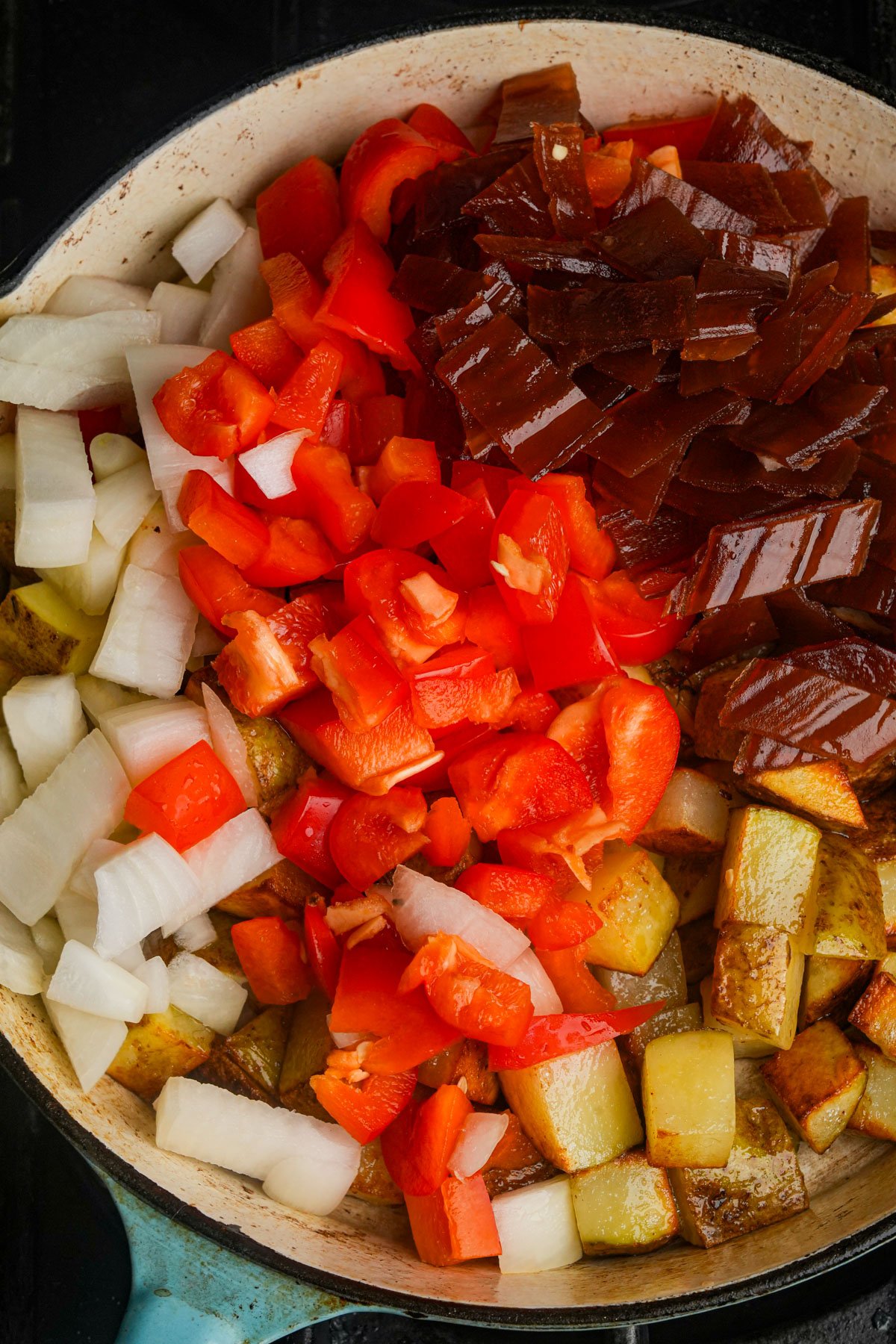 Chopped onions, red bell peppers, potatoes, and a veggie bacon cooking together in a skillet.
