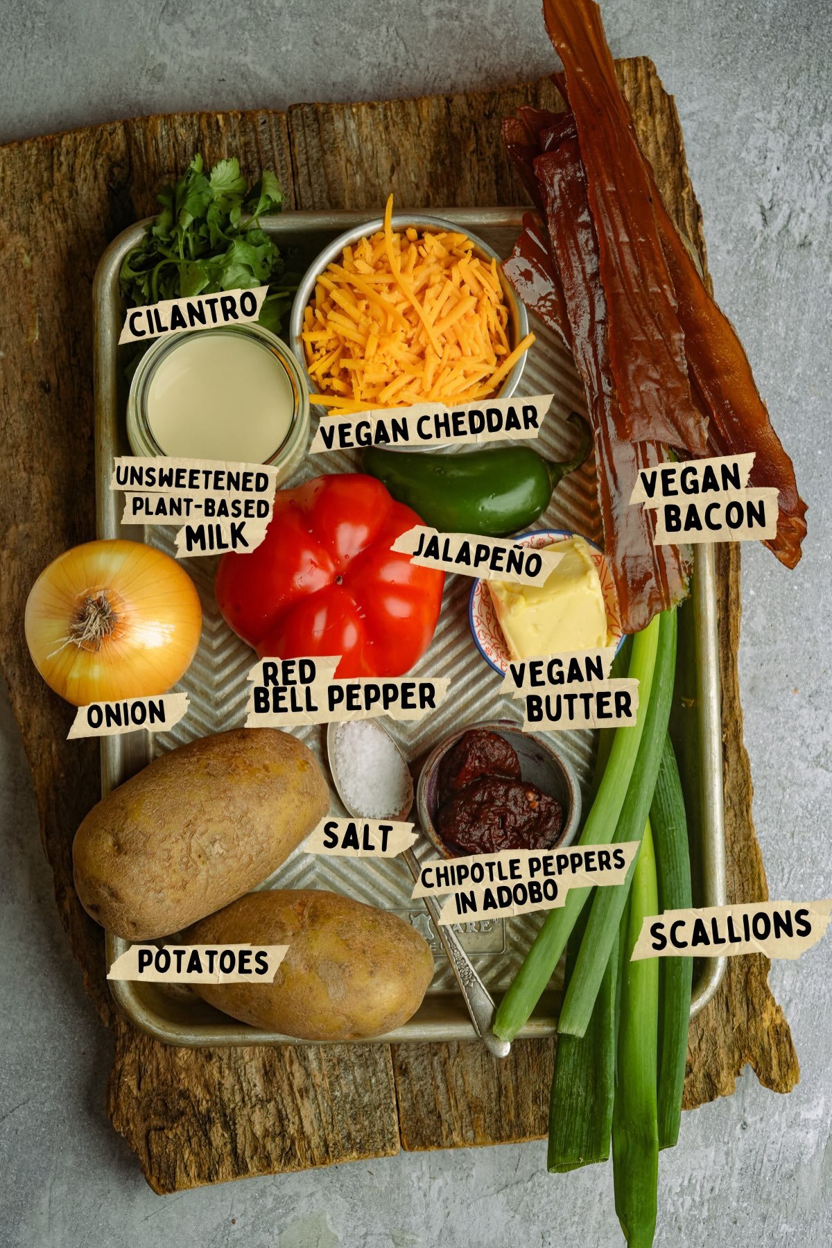 A tray with labeled ingredients for a vegan breakfast hash recipe: cilantro, vegan cheddar, vegan bacon, unsweetened plant-based milk, onion, red bell pepper, jalapeño, vegan butter, chipotle peppers in adobo, scallions, potatoes, salt.