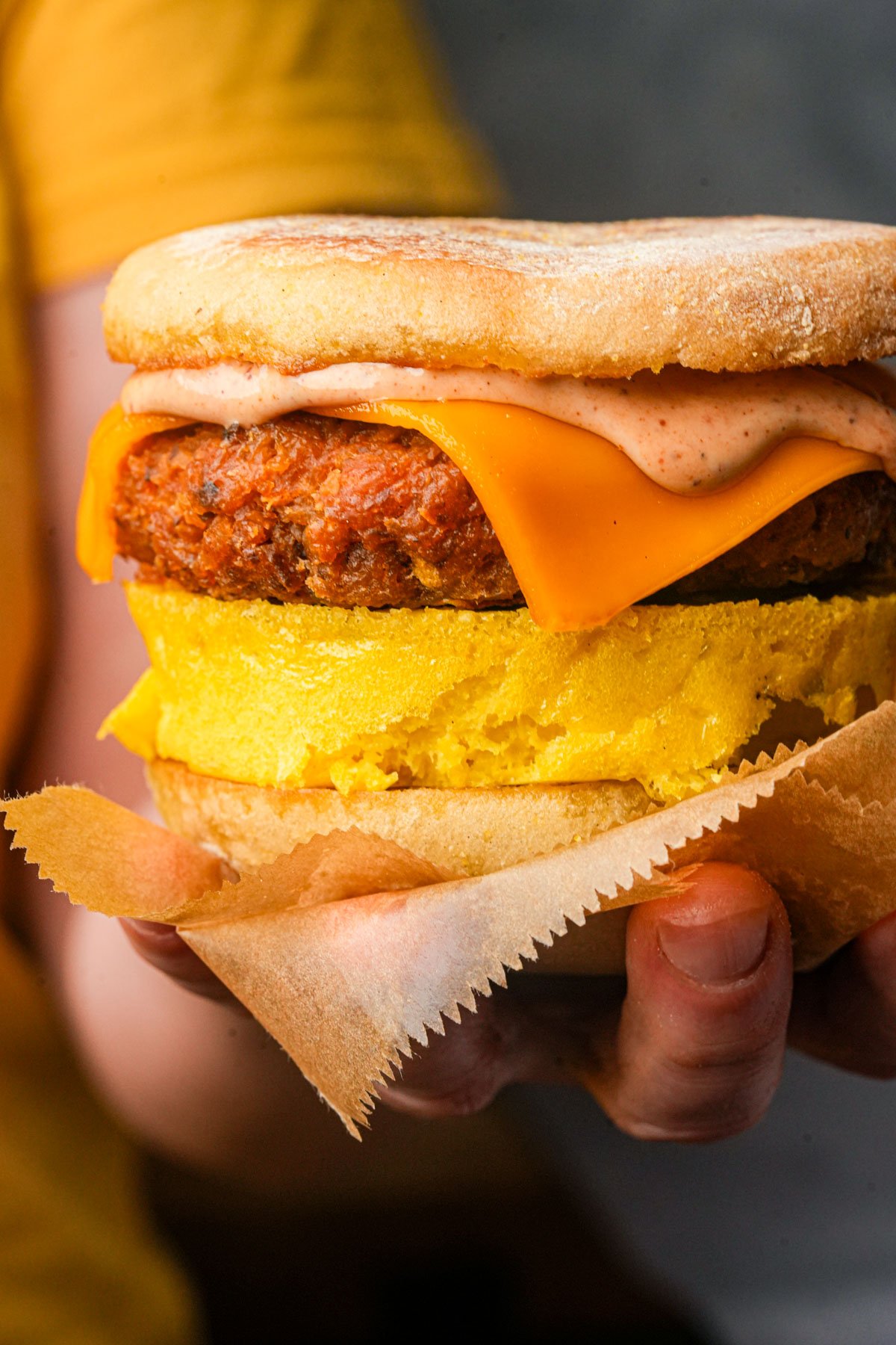A hand holds a vegan breakfast sandwich with an English muffin, Just Egg patty, vegan breakfast sausage patty, cheddar cheese, and sauce.