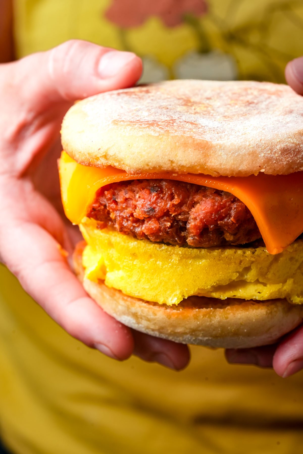A hand holds a vegan breakfast sandwich with a slice of cheddar cheese, a plant-based sausage patty, and a mung bean egg patty.