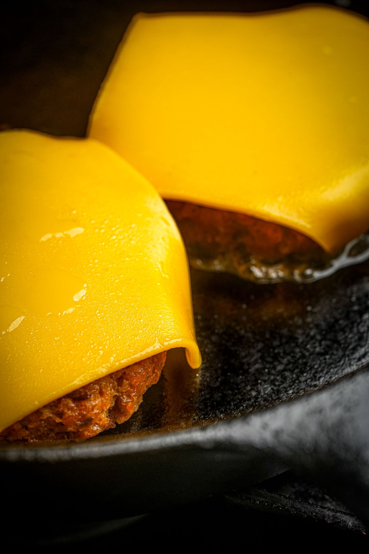 Two vegan breakfast sausage patties topped with slices of yellow cheese are cooking in a black cast iron skillet.