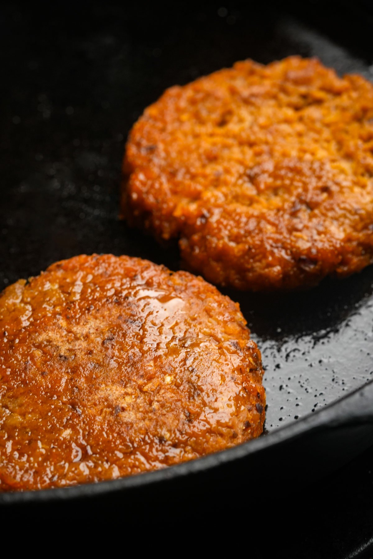 Two plant-based breakfast sausage patties cooking in a black skillet, with visible oil and a browned, textured surface.