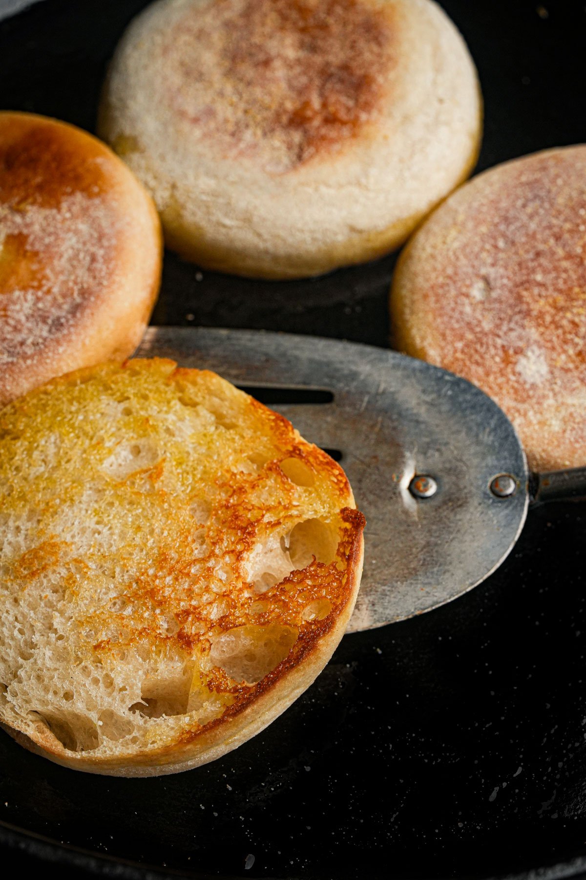 Four English muffins are being toasted in a pan, with one half resting on a metal spatula, showing a golden, crispy surface.