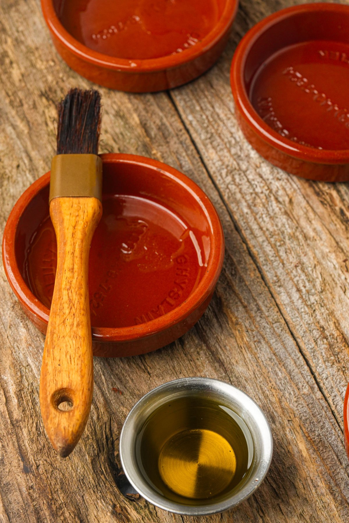 A wooden-handled pastry brush rests on a round brown ceramic dish, with another dish and a small metal cup of oil nearby on a wooden surface.
