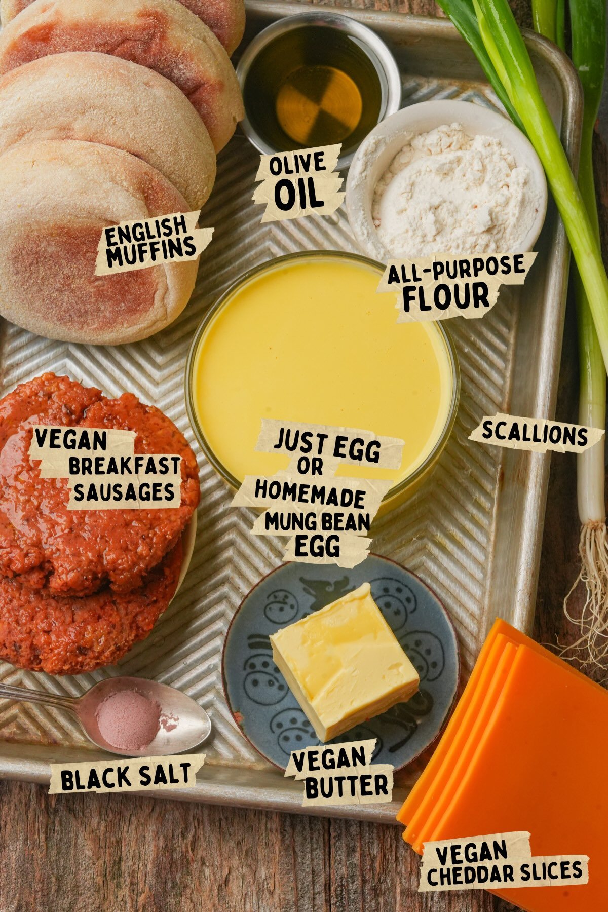 A tray with labeled ingredients for a vegan breakfast sandwich recipe: English muffins, olive oil, all-purpose flour, scallions, vegan breakfast sausages, vegan butter, vegan cheddar slices, black salt, and Just Egg.