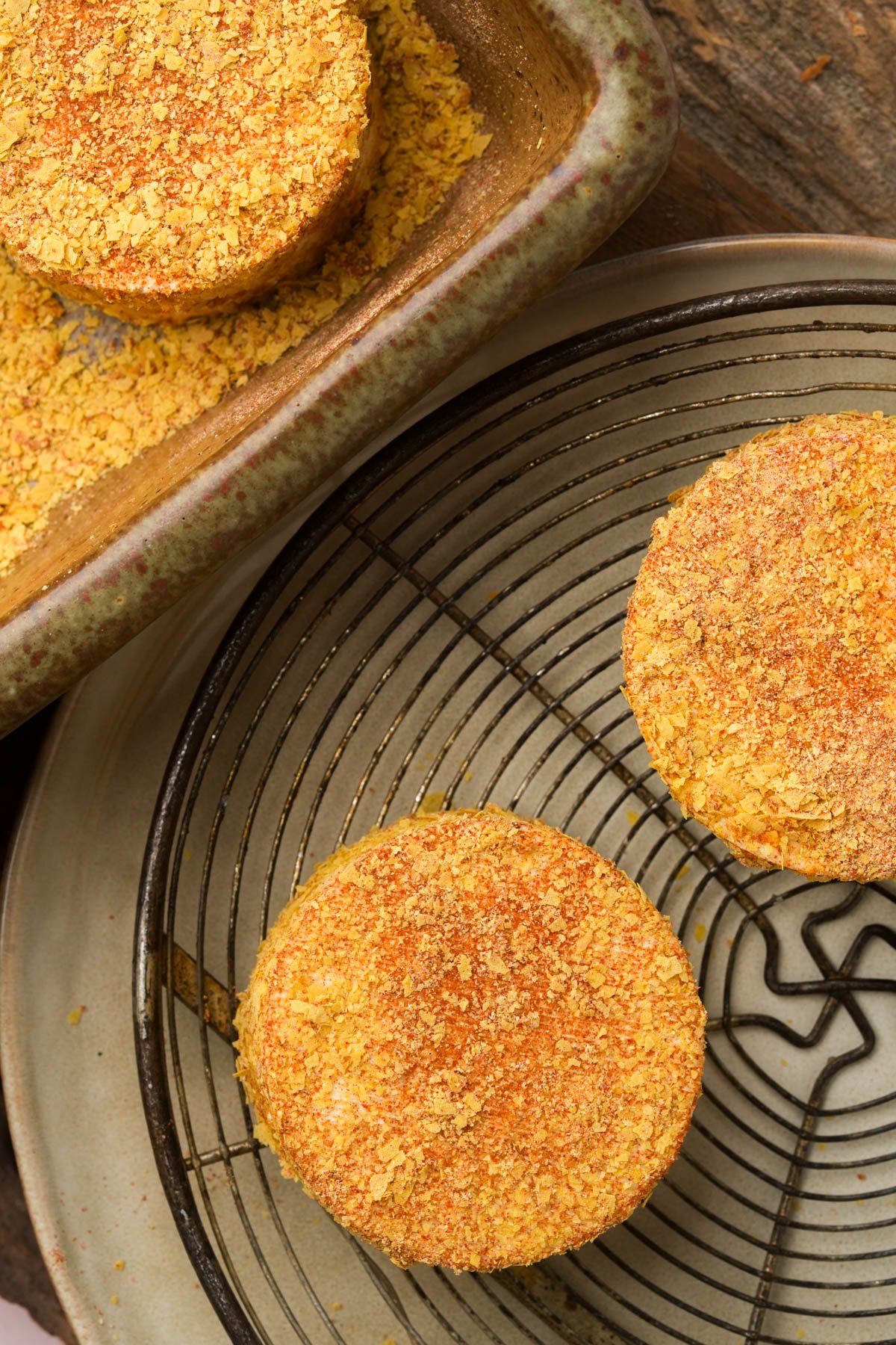 Three round, seasoned tofu patties sit on a wire rack and in a baking dish, coated with a yellow crumb mixture on a rustic wooden surface.