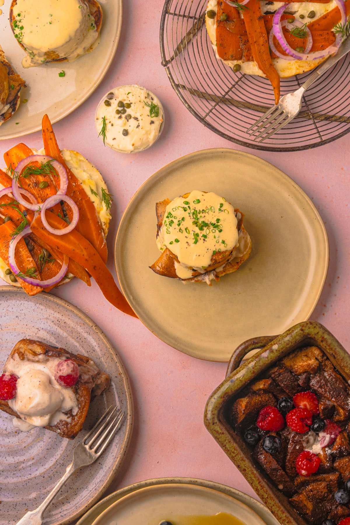 Assorted brunch dishes including vegan eggs benedict, toast with vegan smoked salmon and carrots, bread pudding with berries, and a cinnamon roll with icing on various plates on a pink surface.