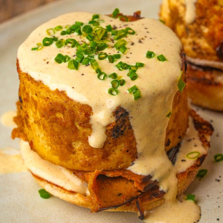 A thick serving of vegan eggs benedict with creamy sauce and chopped chives, served on an English muffin with a slice of roasted vegetable.