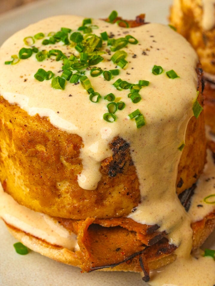 A thick serving of vegan eggs benedict with creamy sauce and chopped chives, served on an English muffin with a slice of roasted vegetable.