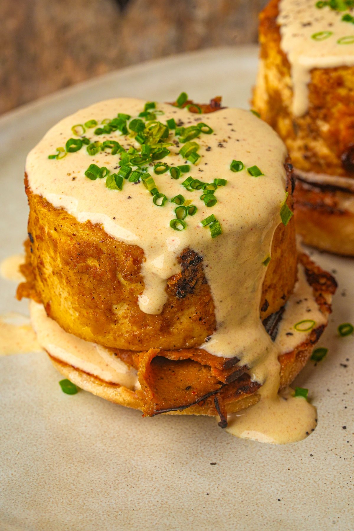 Two thick servings of vegan eggs benedict topped with creamy sauce and chopped chives, served on a beige plate.