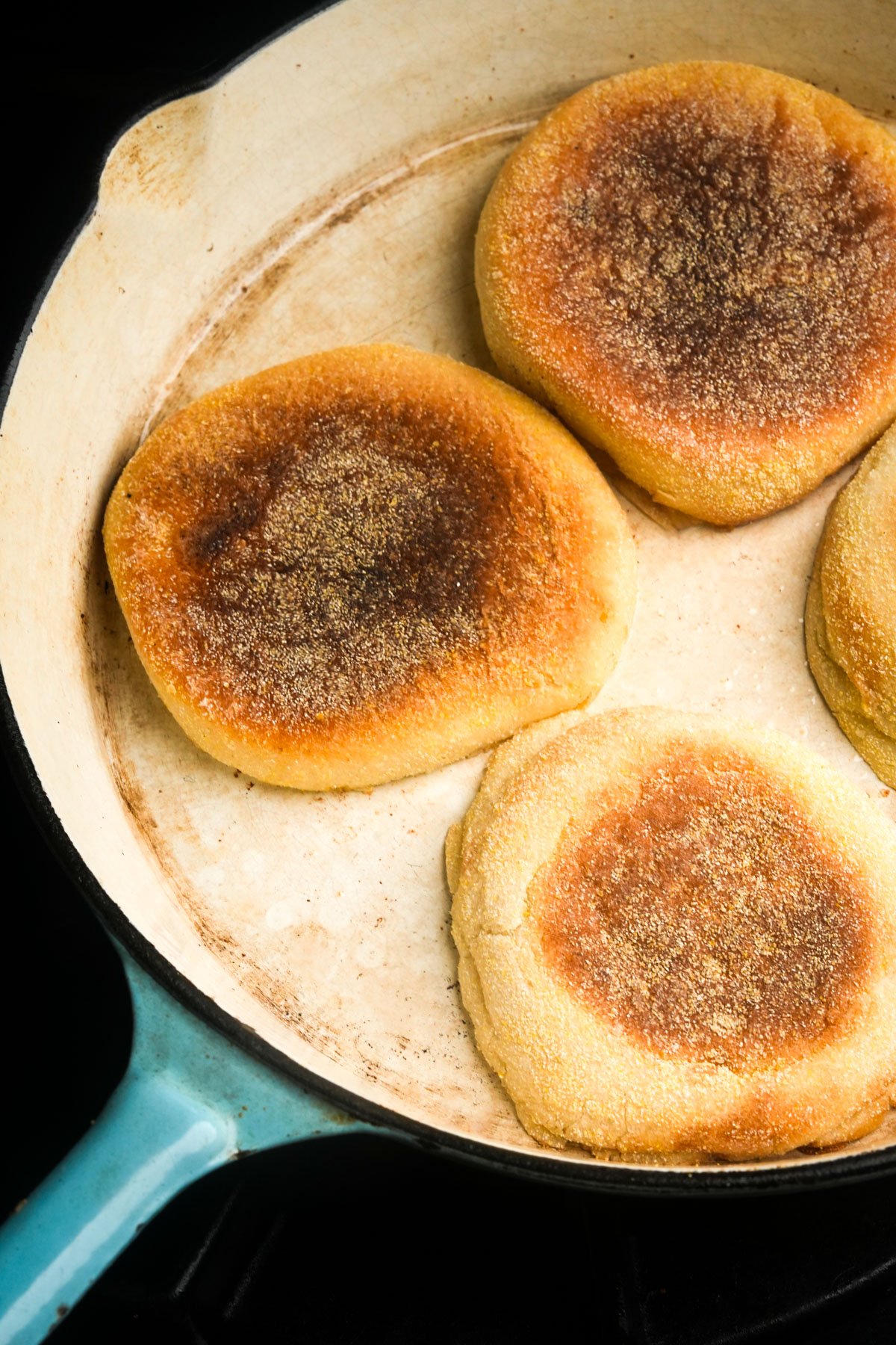Four golden-brown English muffins cooking in a cream-colored skillet with a blue handle.