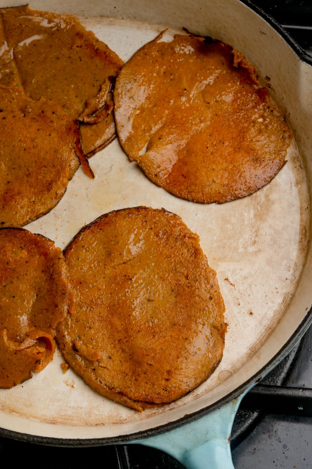 Five browned, round slices of seitan ham are cooking in a large, light-colored skillet on a stovetop.