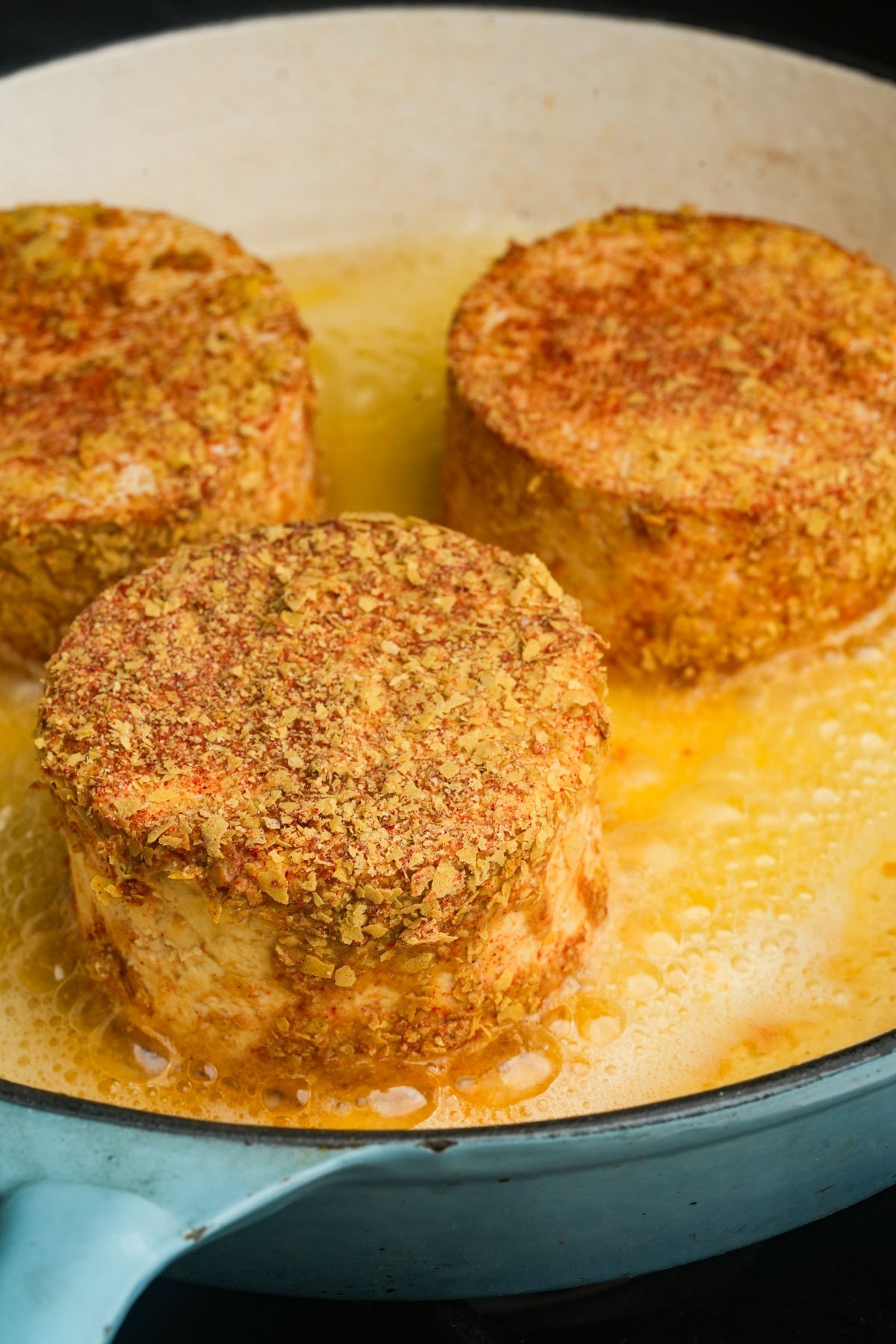Three seasoned, round pieces of tofu are being pan-fried in oil in a light blue skillet.