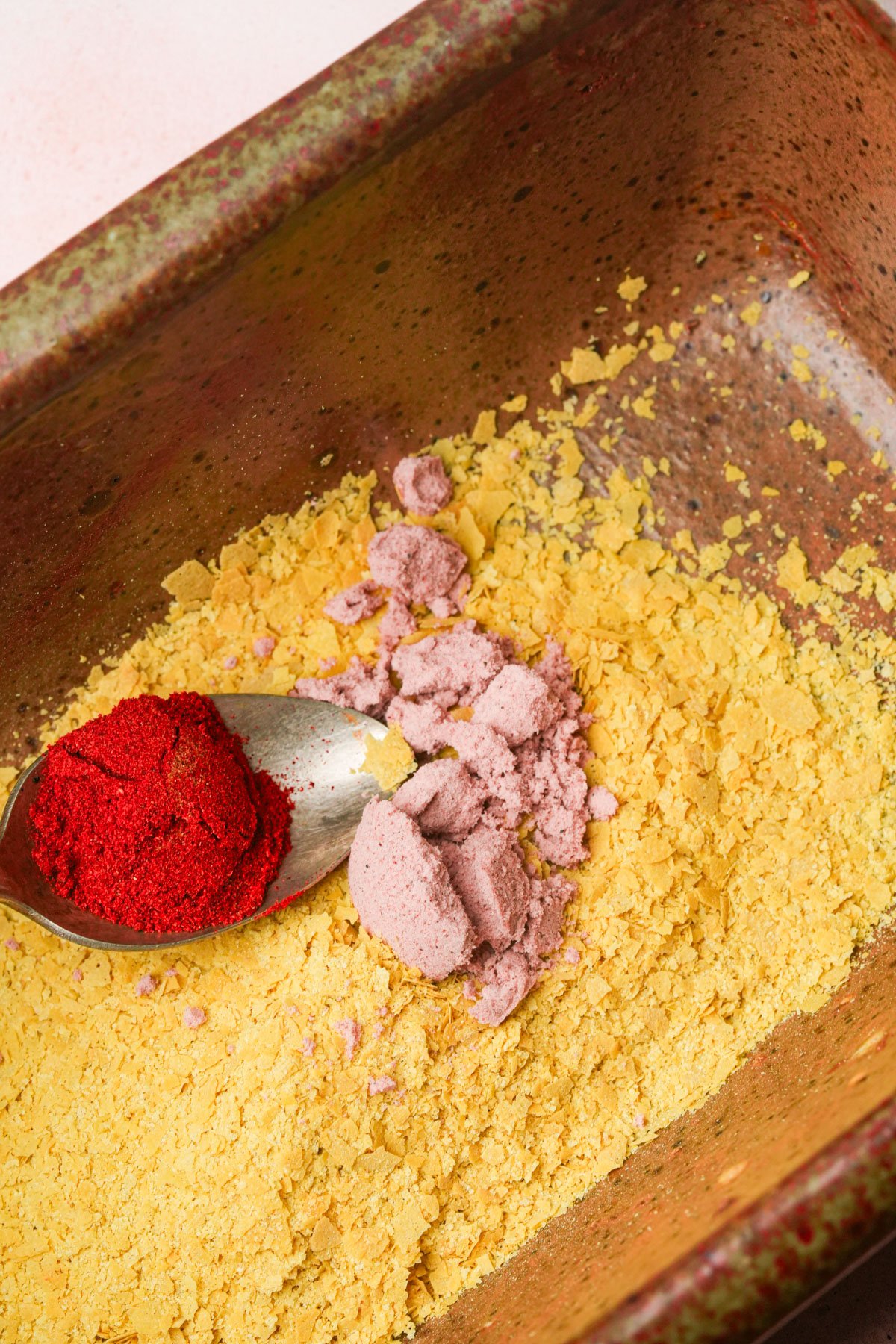 A spoon mixes paprika and kala namak into a dish containing yellow nutritional yeast flakes.