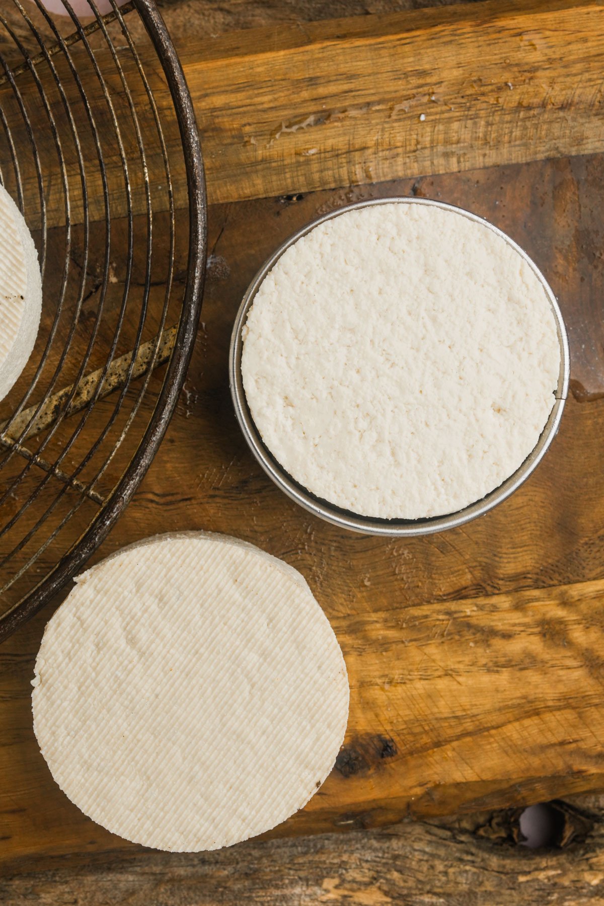 Three round discs of white tofu rest on a wooden surface, with one placed inside a metal ring and another on a wire cooling rack.