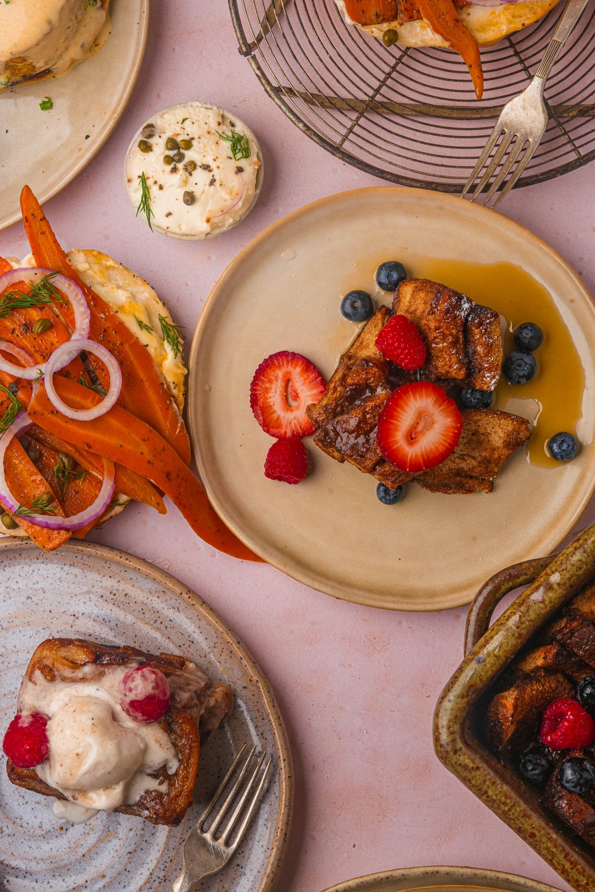 Plates of vegan French toast casserole topped with fresh berries and syrup are arranged alongside a bagel with vegan smoked salmon, vegan cream cheese, and vegan eggs benedict with hollandaise sauce on a pink surface.