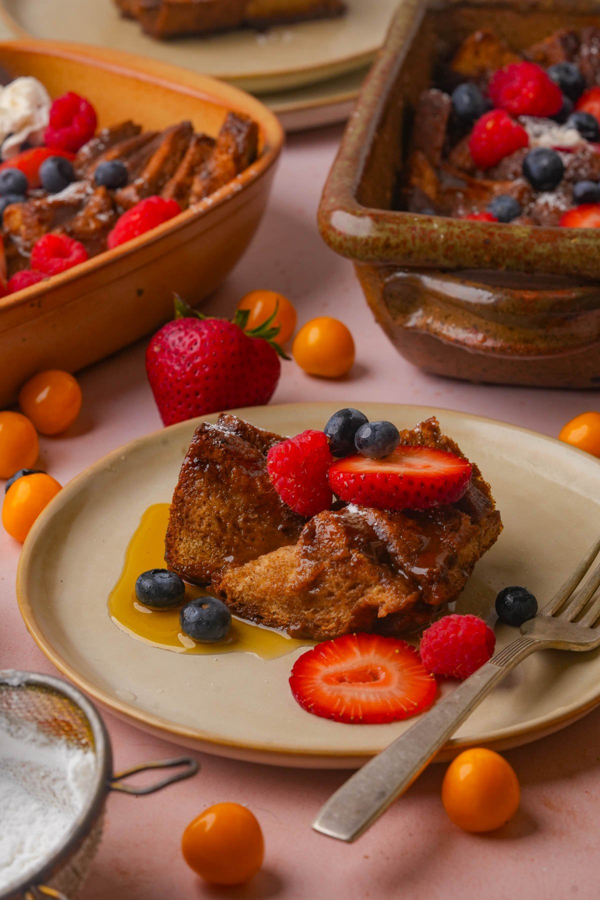 A plate with a slice of vegan baked French toast topped with strawberries, raspberries, blueberries, and syrup. Other serving dishes and fresh fruit are also visible on the table.