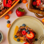 A plate with vegan French toast casserole topped with fresh strawberries, blueberries, and syrup, surrounded by more fruit and baking dishes with additional servings.