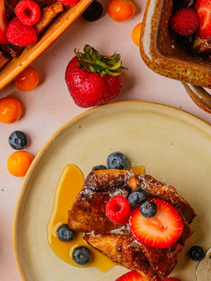 A plate with vegan French toast casserole topped with fresh strawberries, blueberries, and syrup, surrounded by more fruit and baking dishes with additional servings.