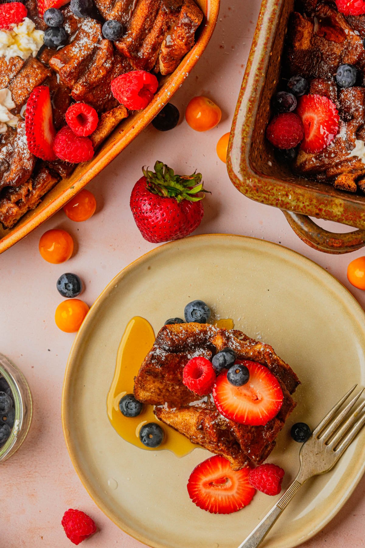 A plate with a slice of vegan French toast casserole topped with strawberries, raspberries, and blueberries, surrounded by dishes of more French toast and scattered berries.