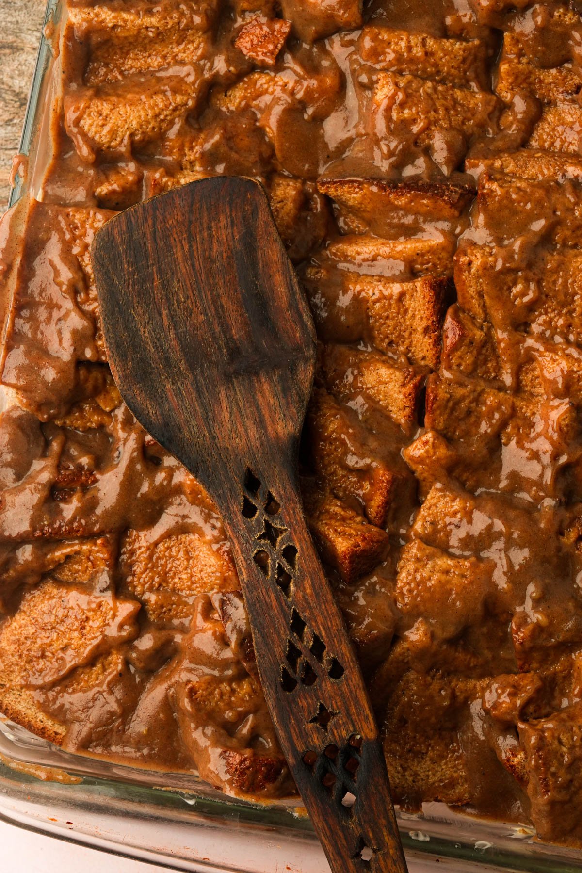 A vegan French toast casserole in a glass dish, topped with brown custard sauce, with a dark wooden spatula resting on top.