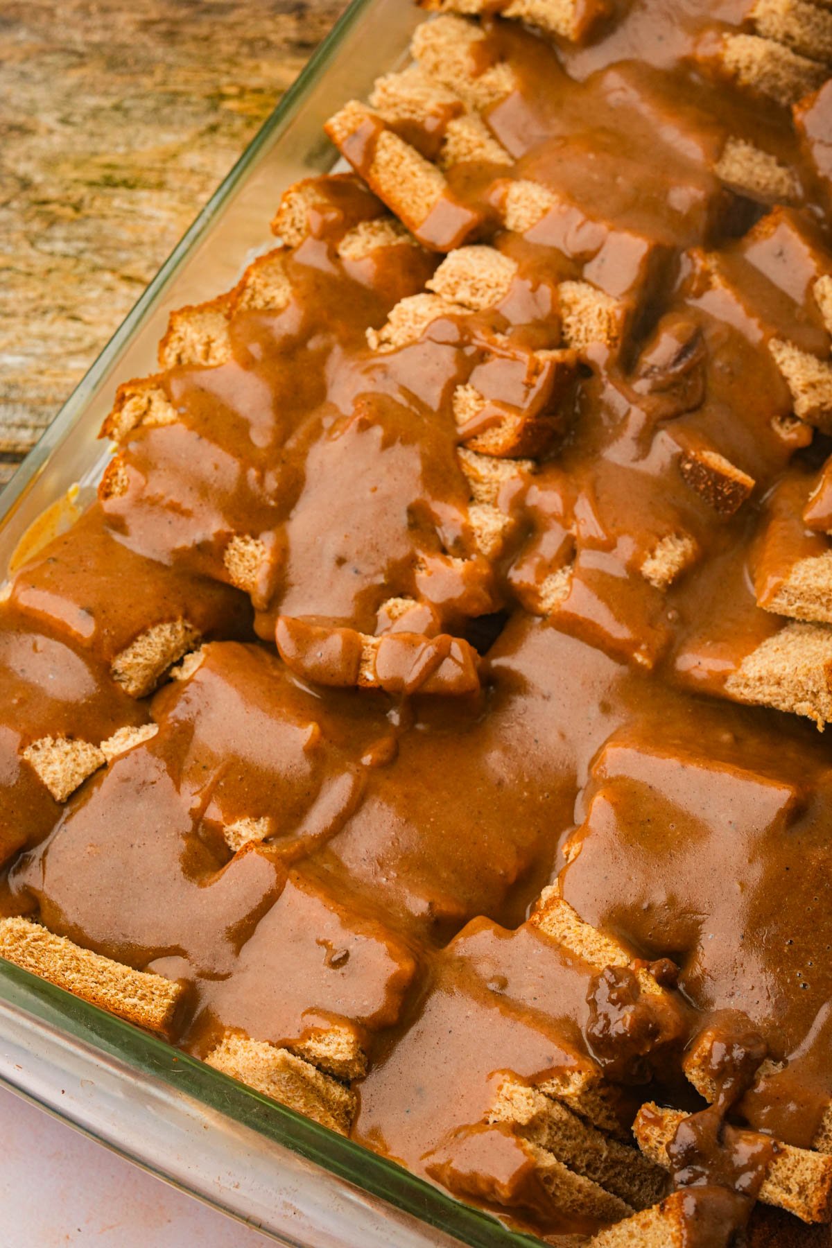 A glass baking dish filled with vegan French toast casserole covered in a thick brown custard sauce, set on a rustic wooden surface.