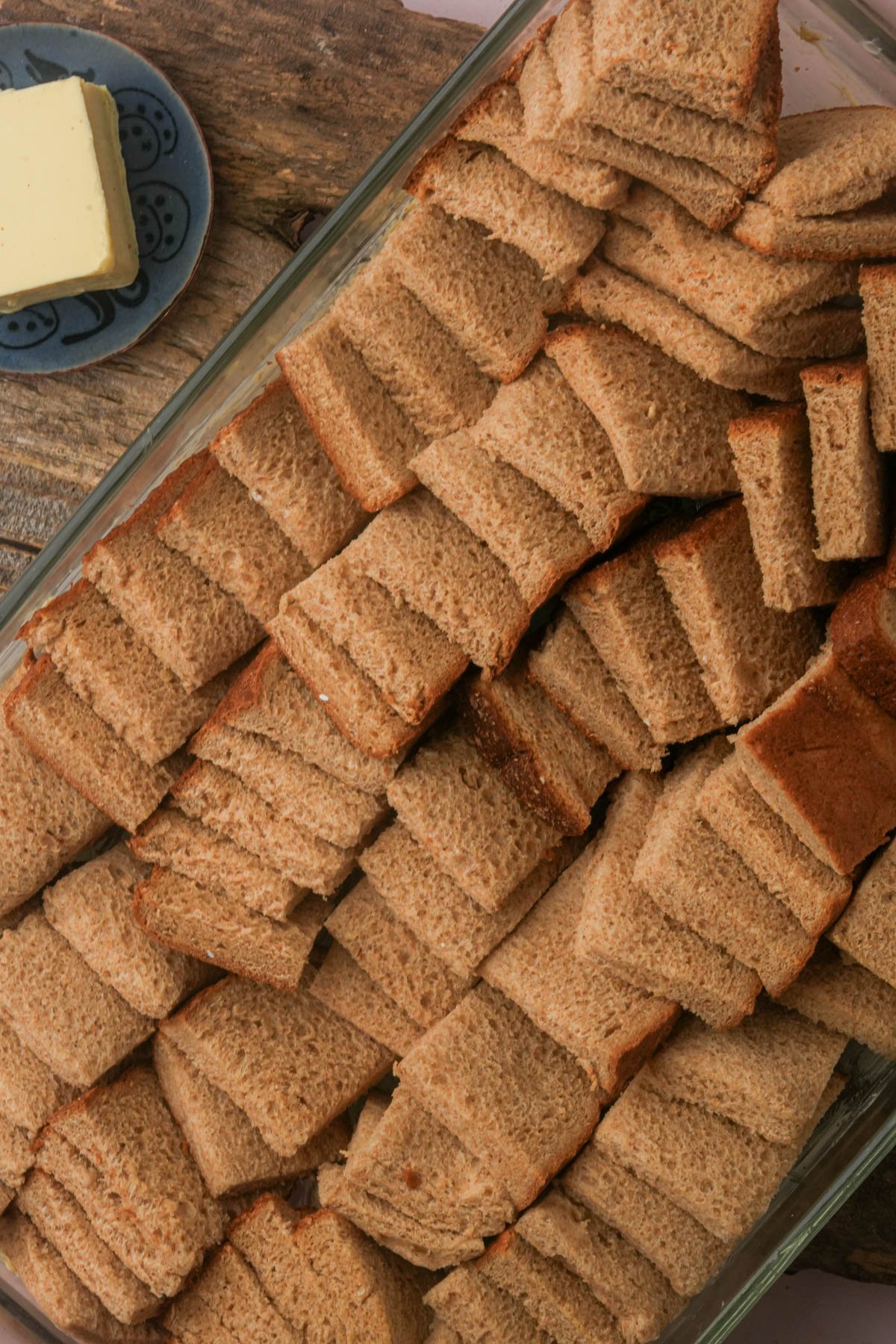 A glass dish filled with neatly stacked pieces of crustless whole wheat bread. A plate with a block of butter is visible nearby on a wooden surface.