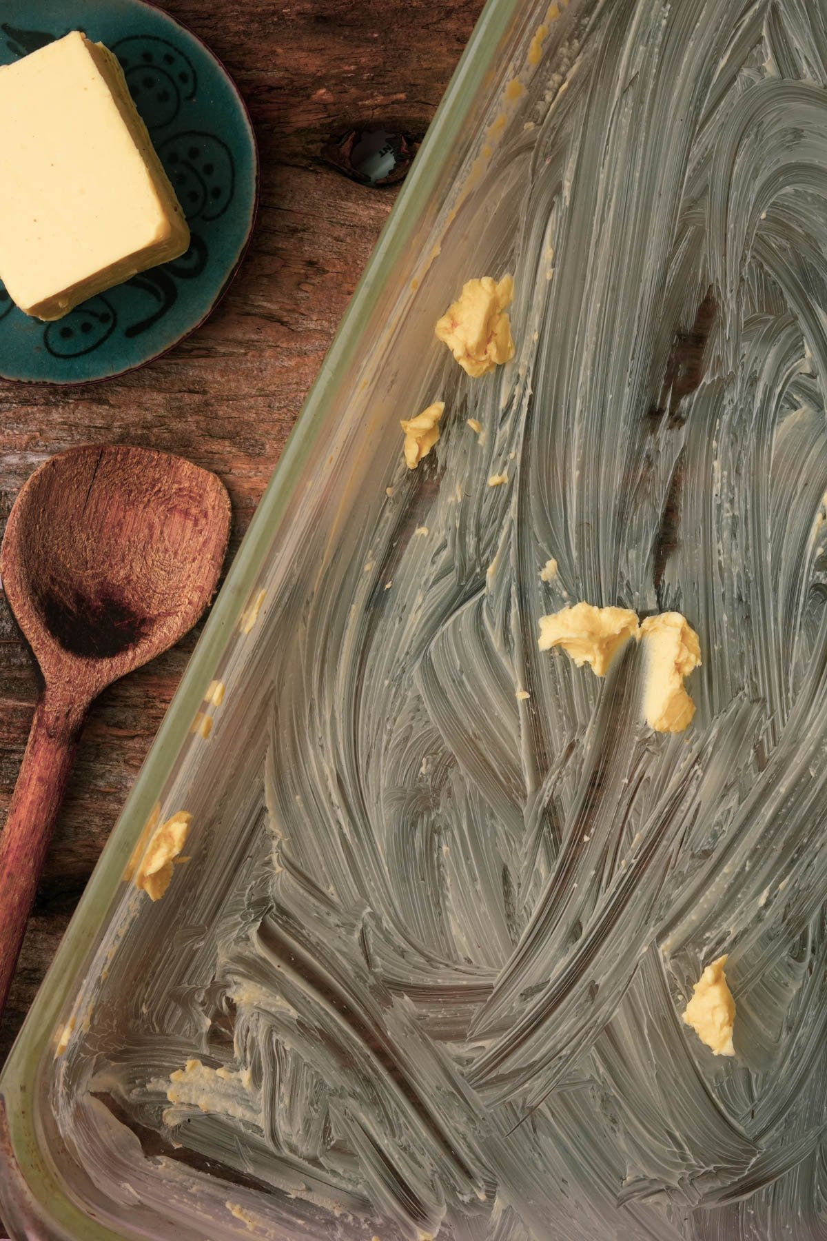 A glass baking dish smeared with vegan butter sits on a wooden surface beside a wooden spoon and a plate holding a block of butter.