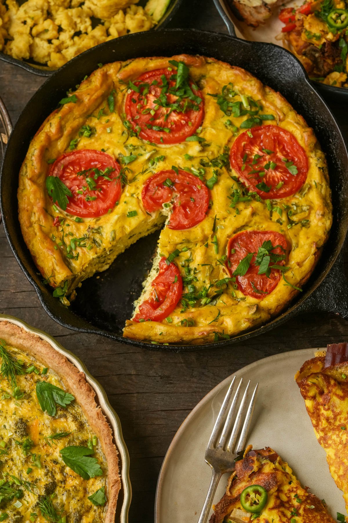 A vegan frittata in a cast iron skillet, topped with sliced tomatoes and herbs, with one slice missing. Other dishes and a plate with a serving are nearby.
