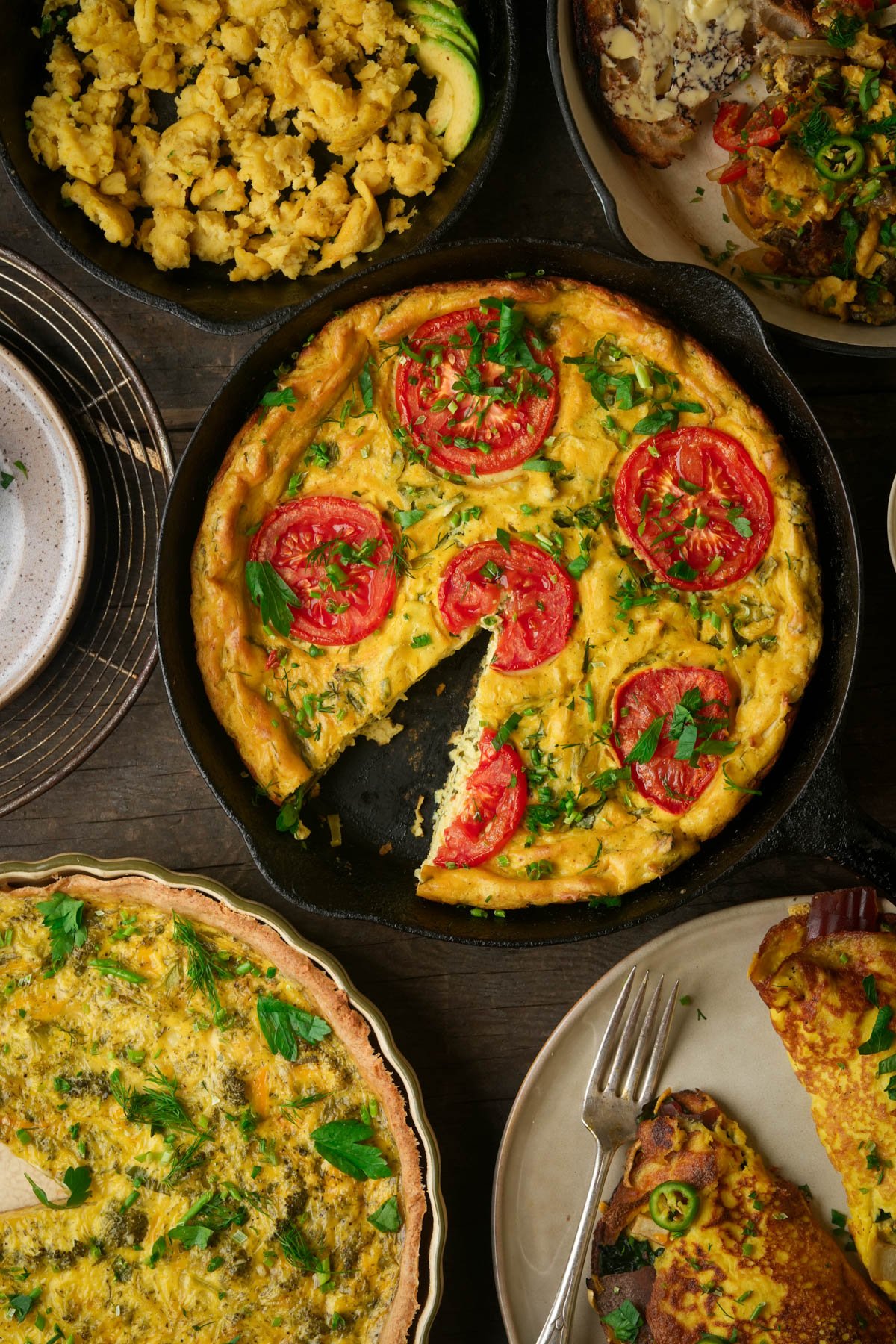 A baked vegan frittata with sliced tomatoes and herbs sits in a skillet, surrounded by plates of scrambled eggs and assorted egg dishes on a wooden table.
