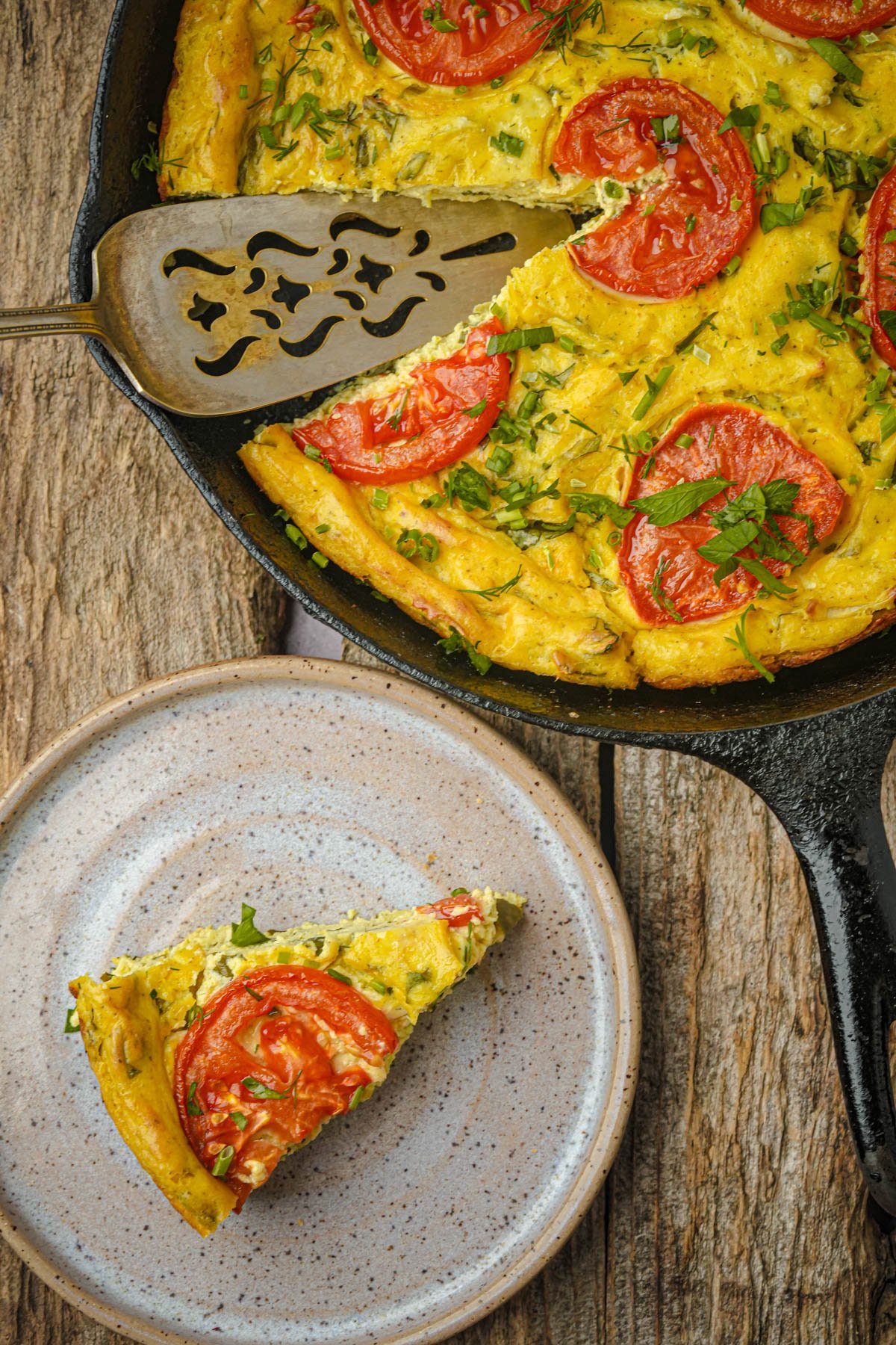 A slice of vegan frittata with tomato and herbs on a plate, next to a cast iron skillet containing the rest of the frittata.