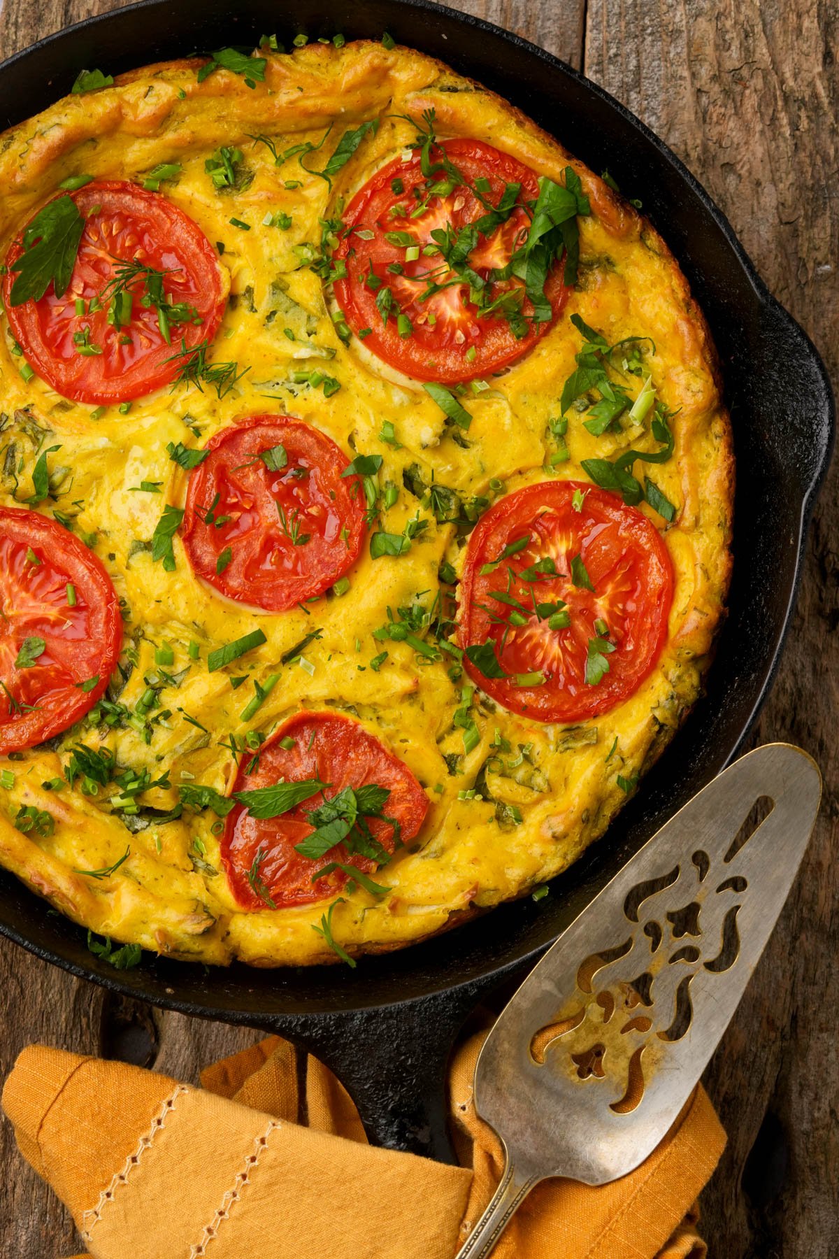 A baked vegan frittata in a cast iron skillet, topped with sliced tomatoes and chopped herbs, with a metal serving spatula and a folded napkin beside it.