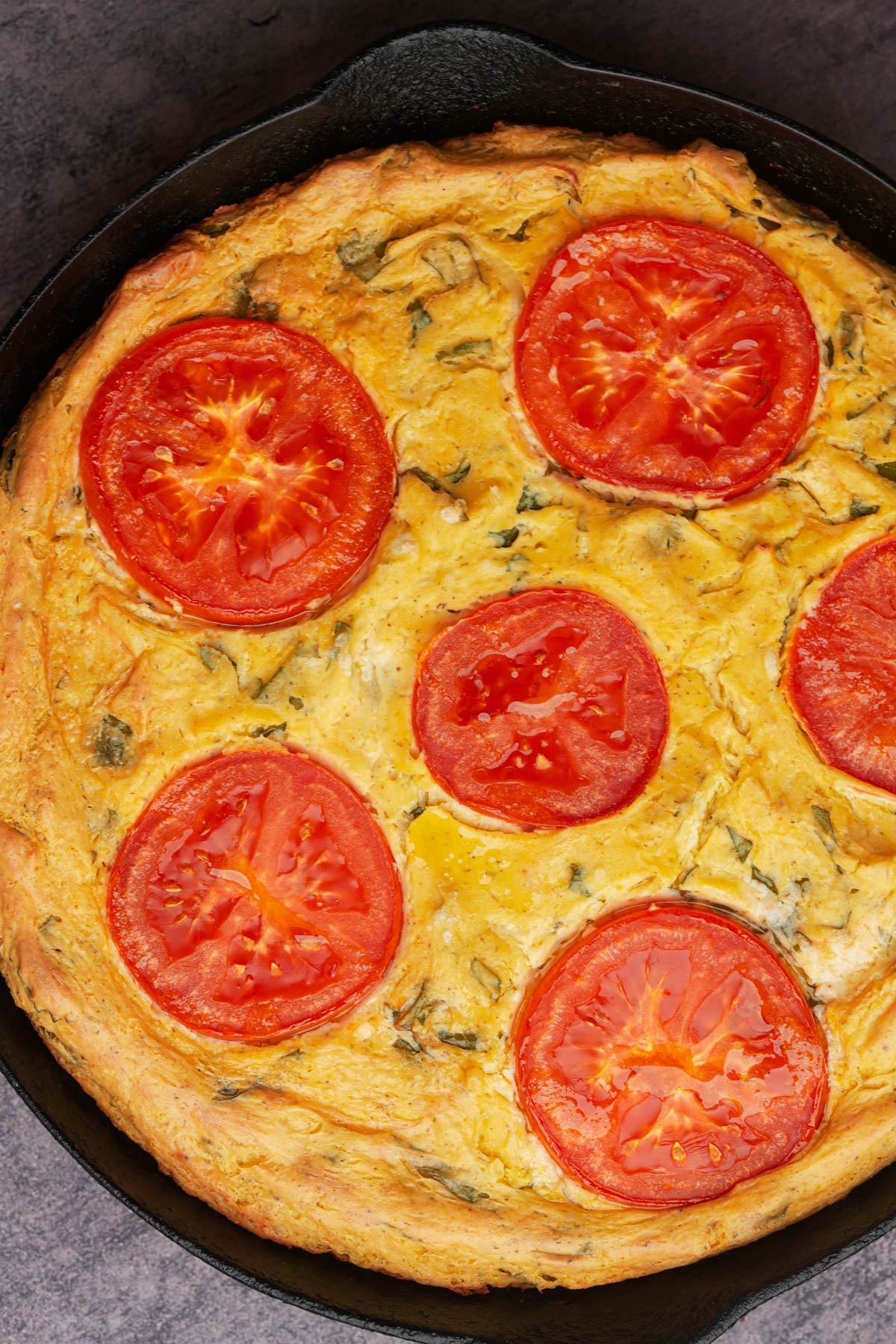 A baked vegan frittata in a skillet, topped with five slices of fresh tomato on a golden, tofu egg base.