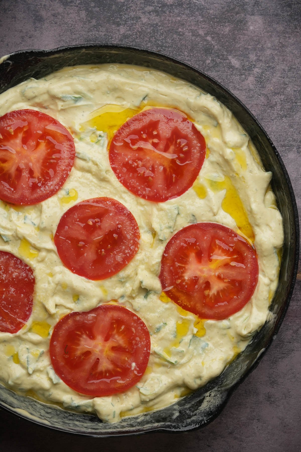 A close-up of an unbaked vegan frittata in a pan, topped with tomato slices set on a textured dark surface.