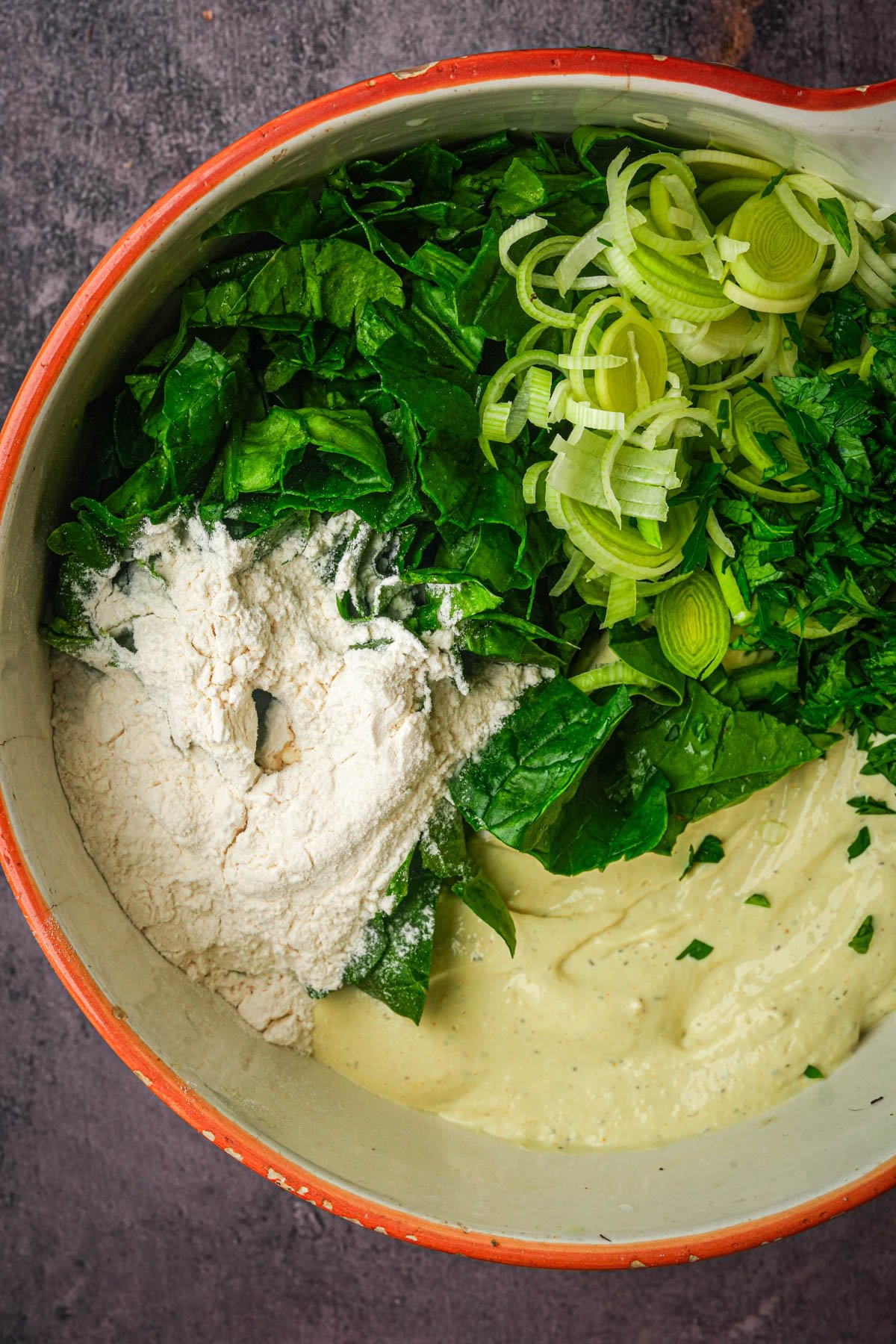 A mixing bowl containing vegan frittata ingredients: chopped spinach, sliced leeks, flour, and a creamy batter, arranged in separate sections before being combined.