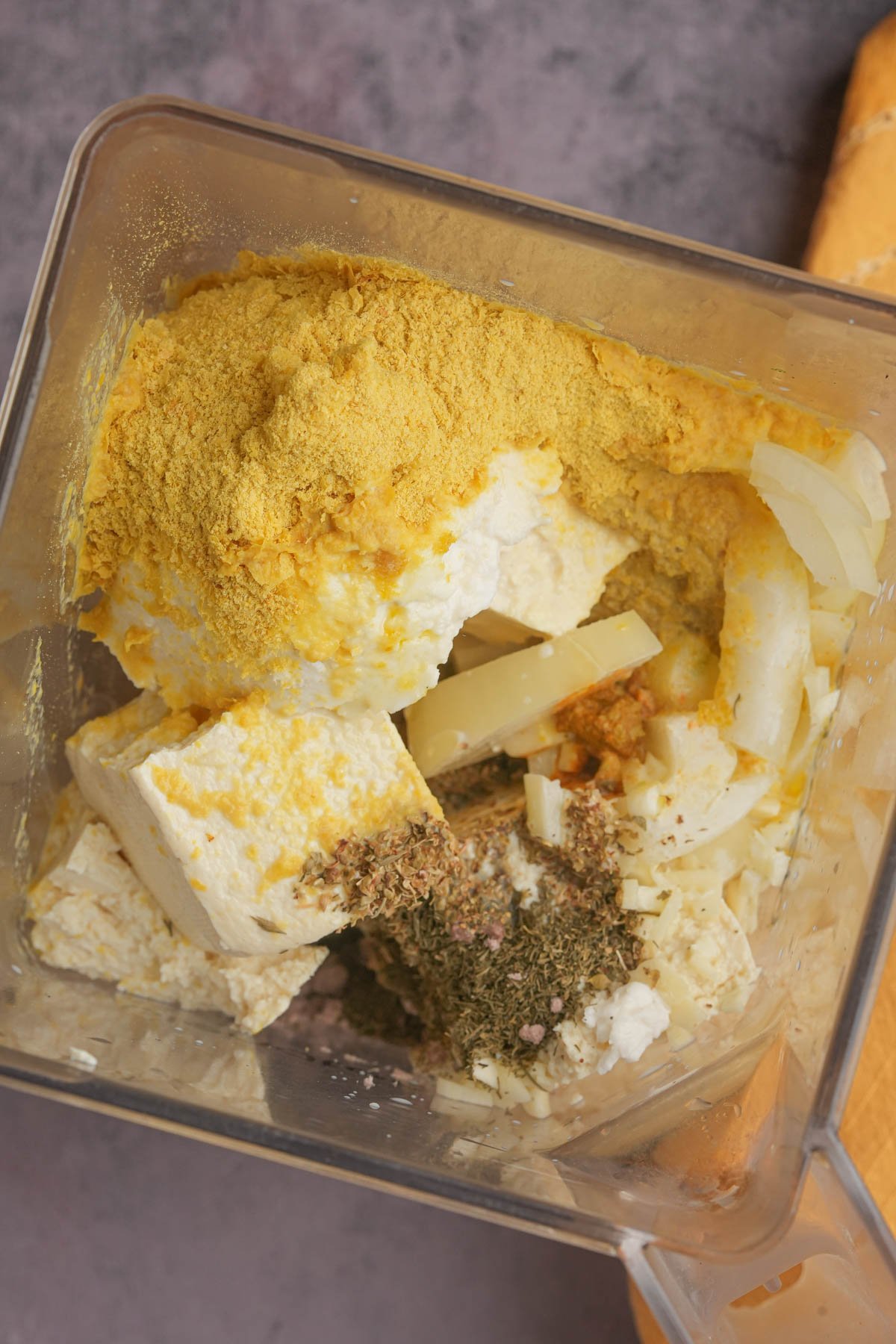 A blender container filled with tofu, nutritional yeast, chopped onion, herbs, and spices, ready to be blended.