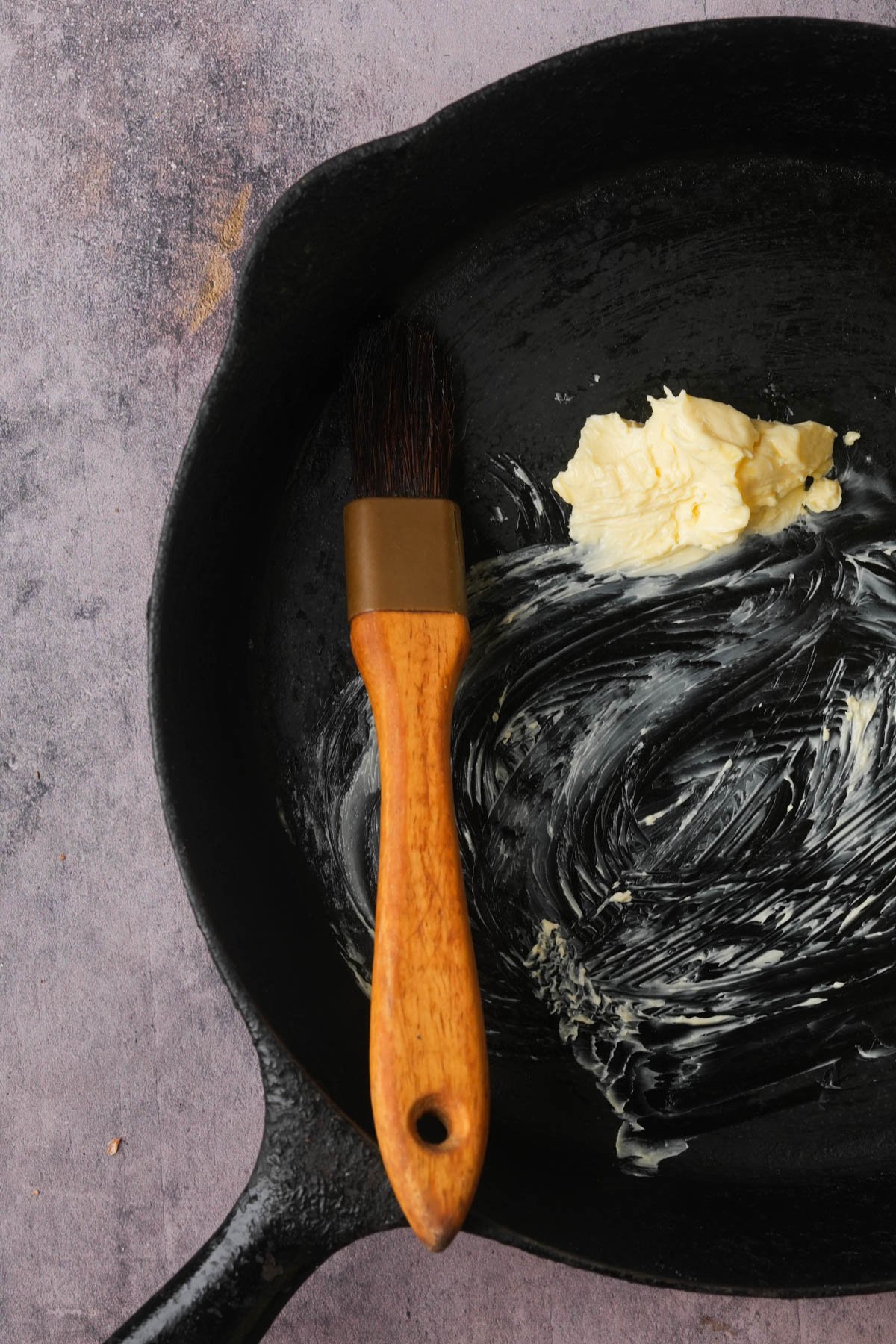 A cast iron skillet with a pat of butter and a wooden-handled brush spreading butter across the surface.