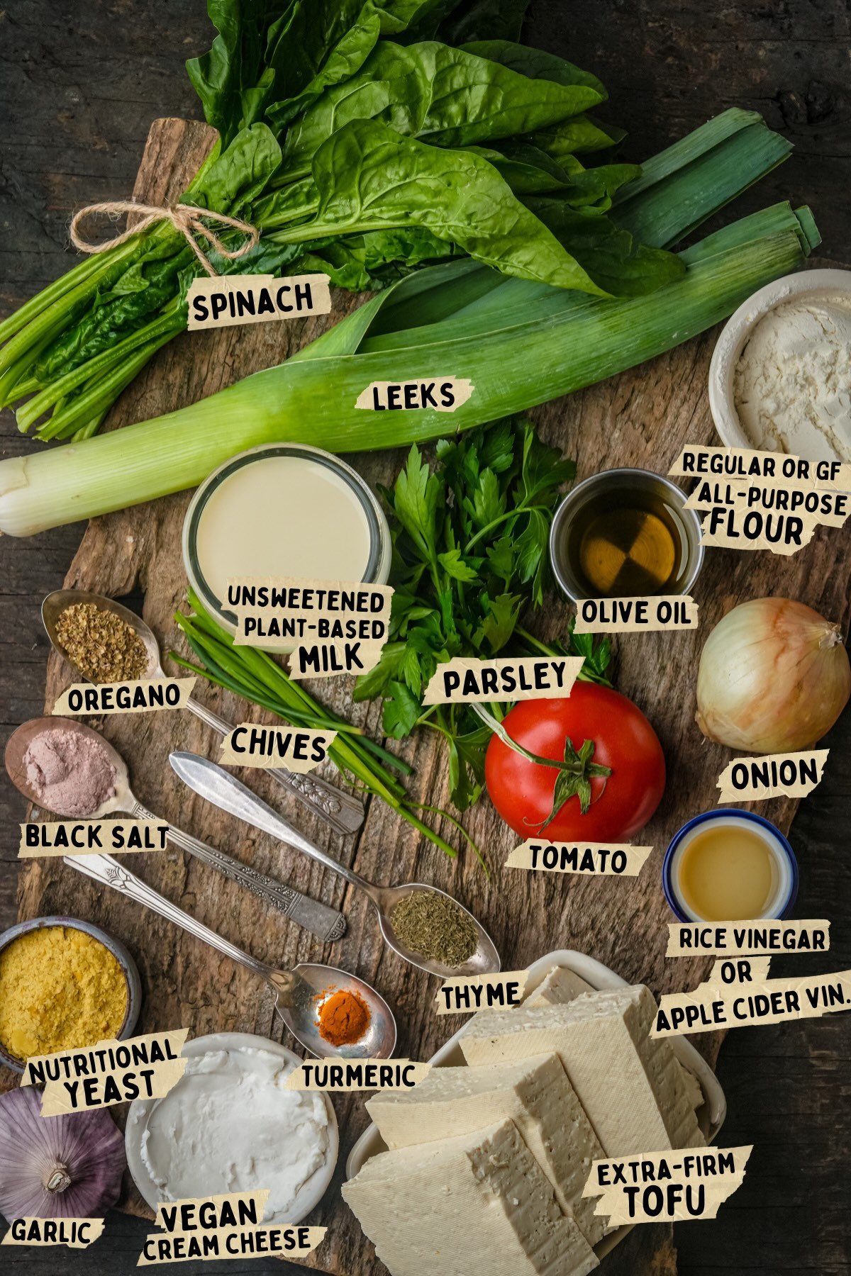 A variety of labeled vegan ingredients for a vegan frittata on a wooden surface, including tofu, spinach, leeks, tomato, onion, spices, herbs, plant-based milk, vegan cream cheese, and flour.