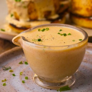A small glass gravy boat filled with creamy vegan hollandaise sauce, garnished with chopped chives and black pepper, sits on a ceramic plate.
