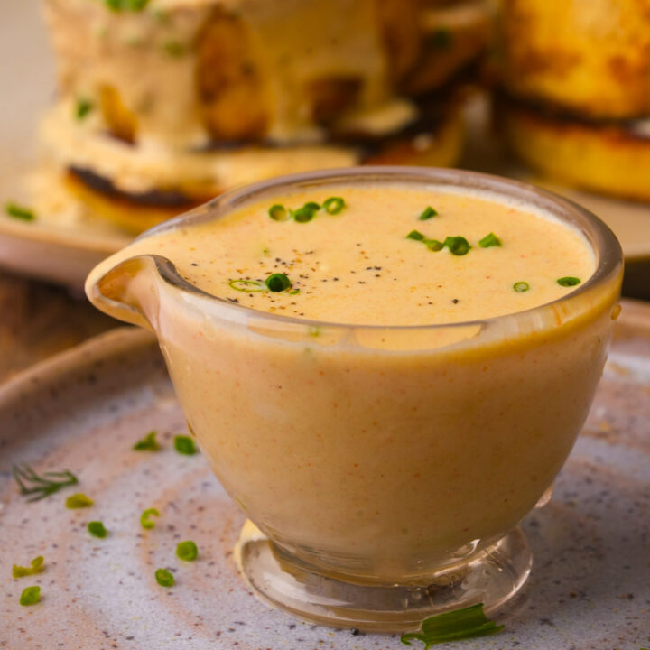 A small glass gravy boat filled with creamy vegan hollandaise sauce, garnished with chopped chives and black pepper, sits on a ceramic plate.