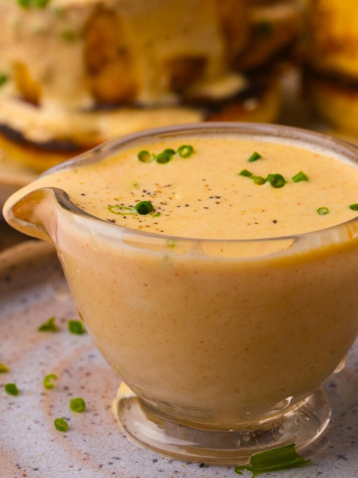 A small glass gravy boat filled with creamy vegan hollandaise sauce, garnished with chopped chives and black pepper, sits on a ceramic plate.