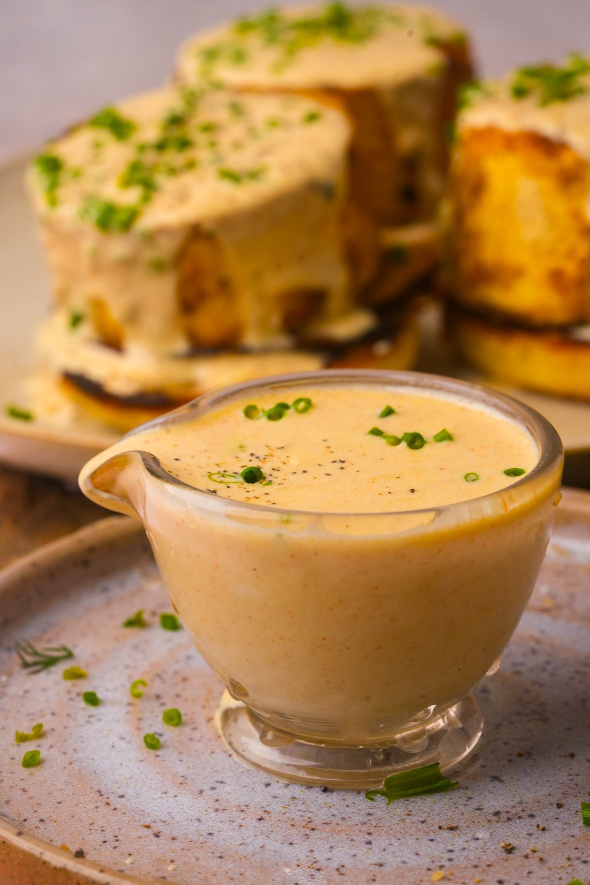A glass gravy boat filled with creamy vegan hollandaise sauce garnished with chives sits on a plate, with sauce-covered food in the background.