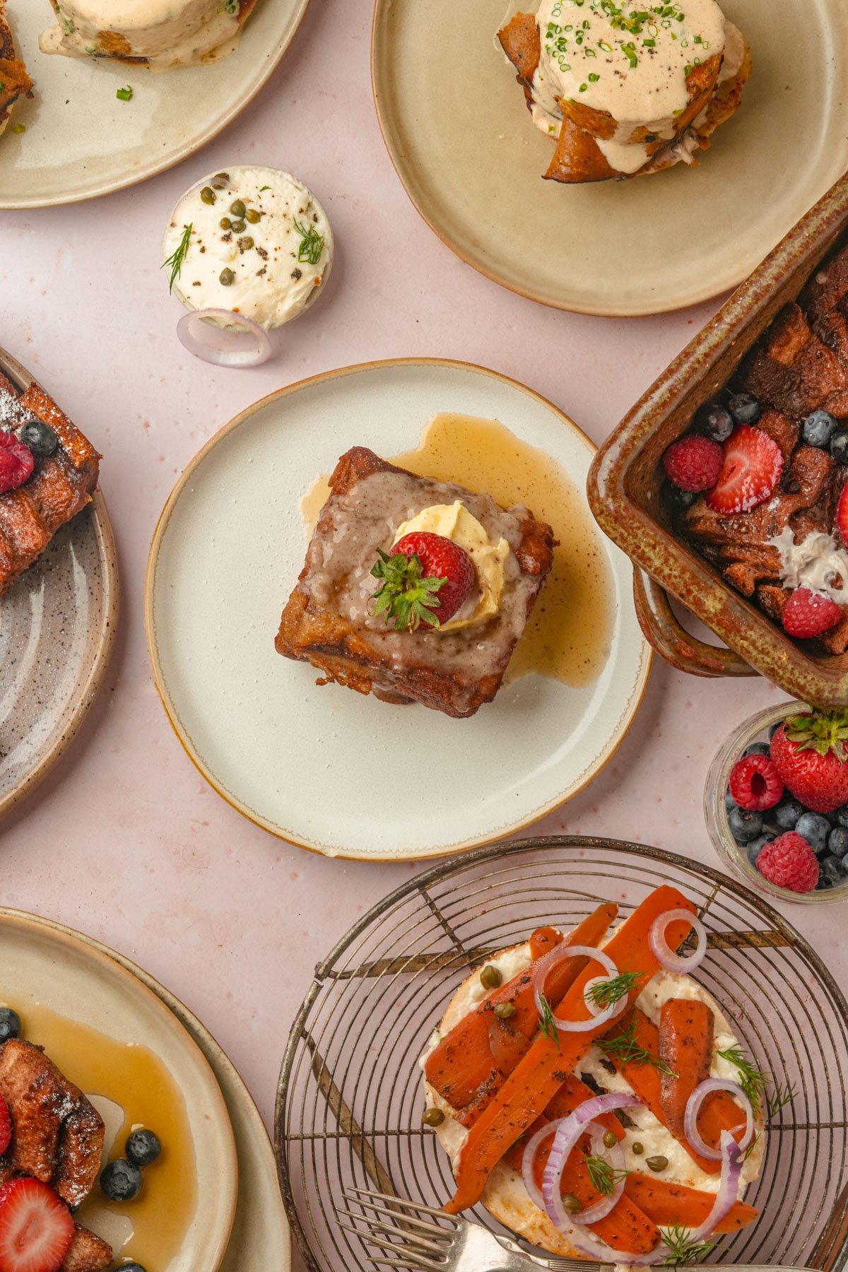 An overhead view of various plated brunch dishes, including Hong Kong french toast, vegan French toast casserole with berries, whipped cream, and garnishes, arranged on a light pink surface.