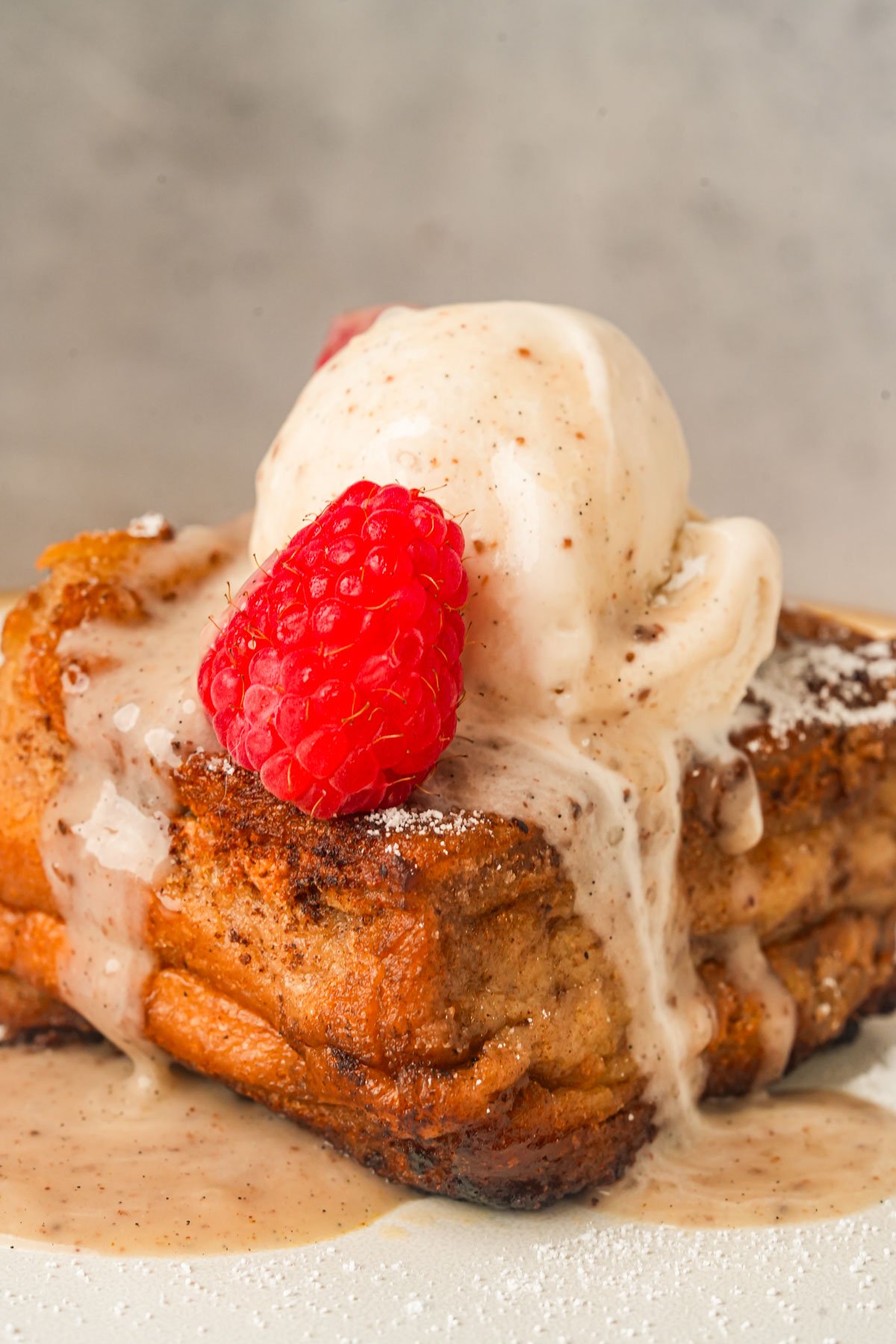 A slice of Hong Kong french toast topped with a scoop of vanilla ice cream, a fresh raspberry, and a drizzle of sauce on a white plate.