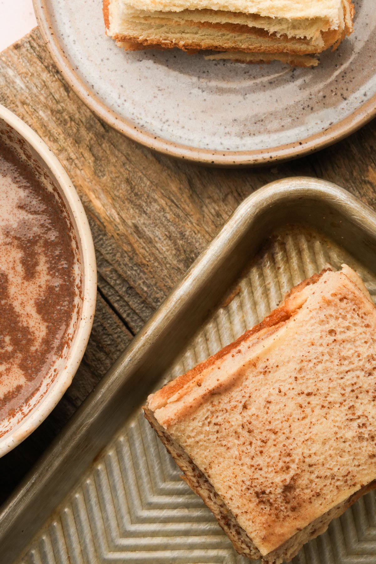 A dredged, uncooked piece of Hong Kong french toast on a baking tray, with a plate of more toast and a bowl of batter on a rustic wooden table.