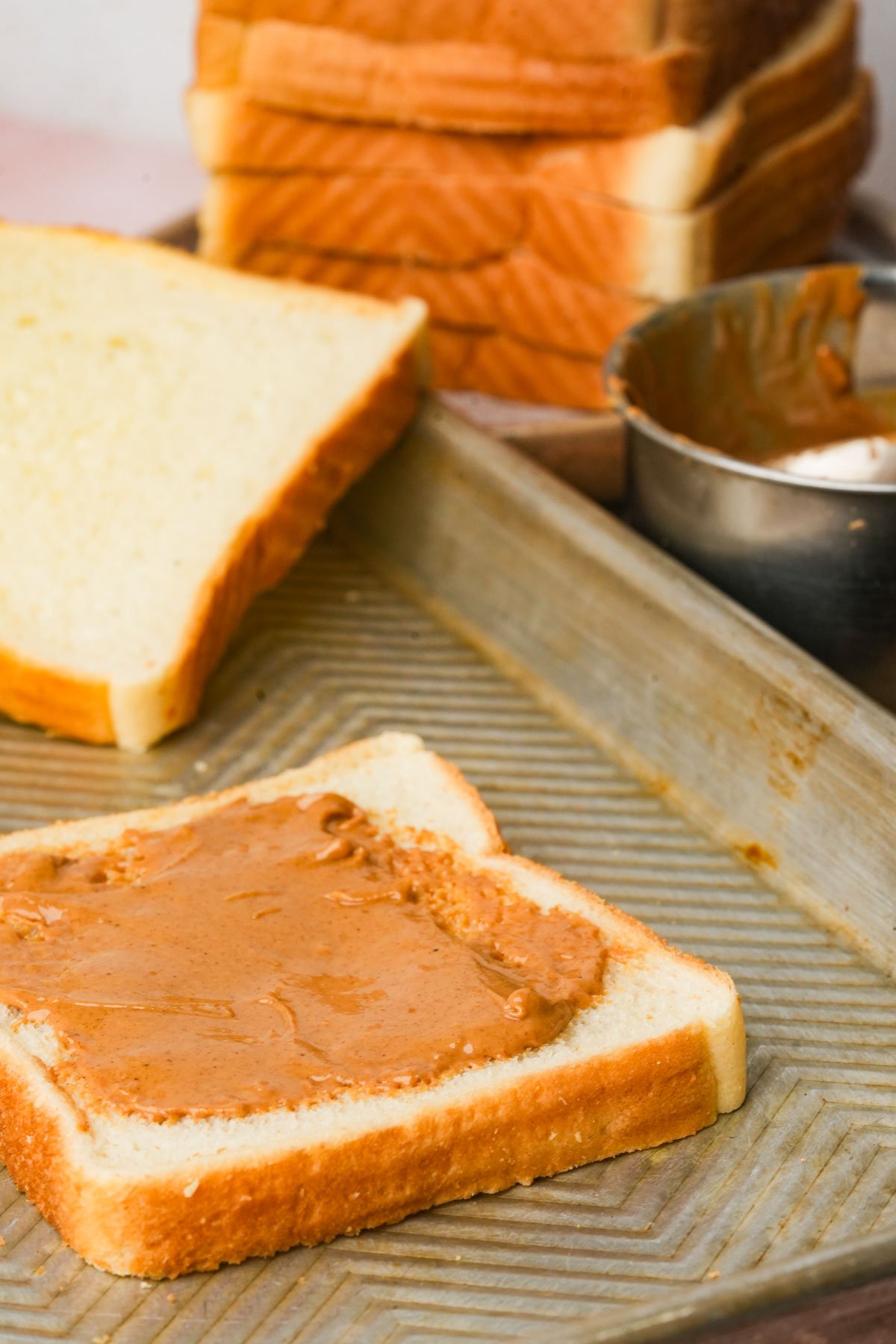 A slice of white bread with peanut butter spread on it sits on a baking sheet, with a fully assembled stack of Hong Kong french toast and a metal bowl in the background.