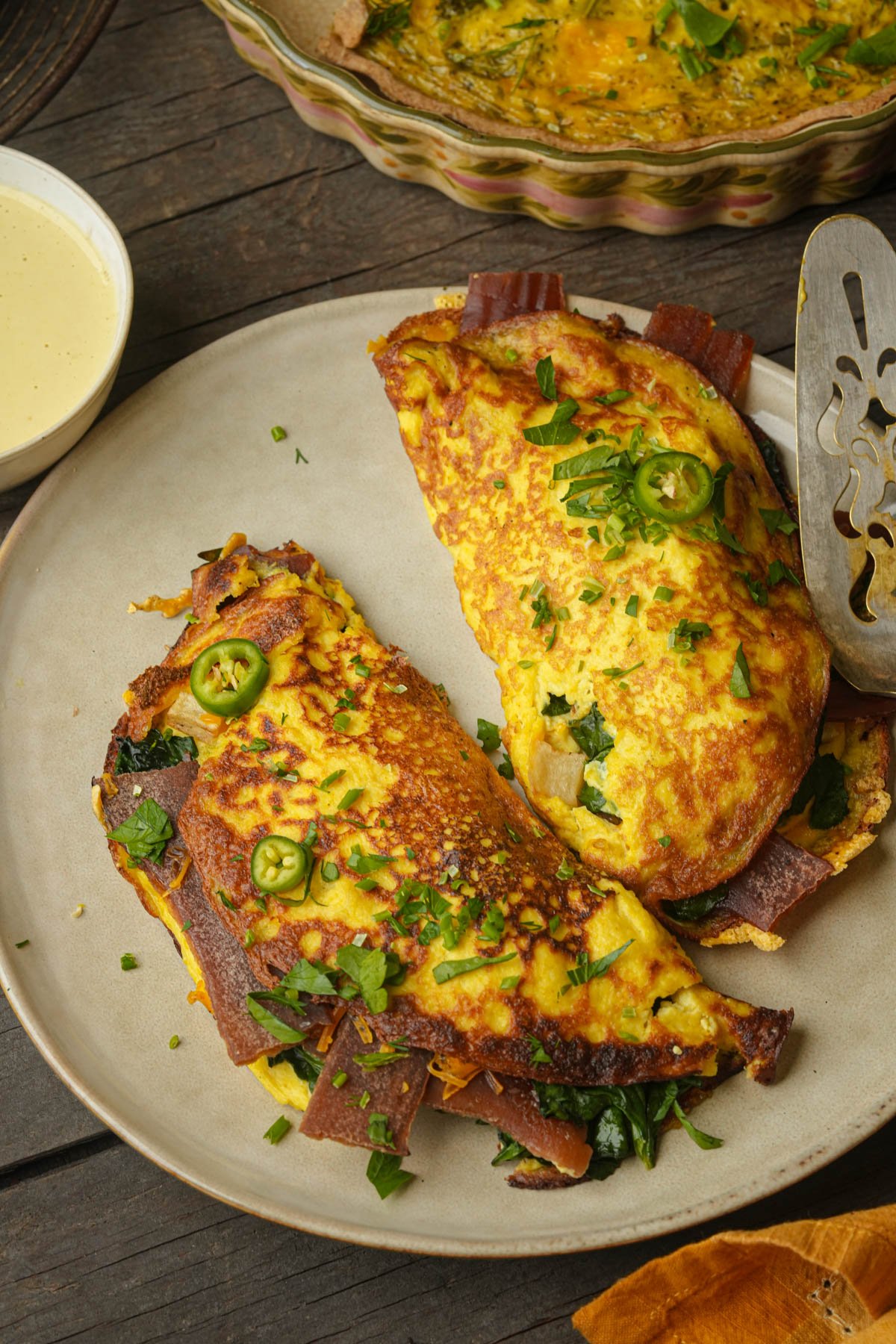 A plate with a folded vegan omelette filled with greens and veggie bacon, garnished with sliced jalapeños and chopped herbs, sits next to a bowl of sauce and a serving utensil.